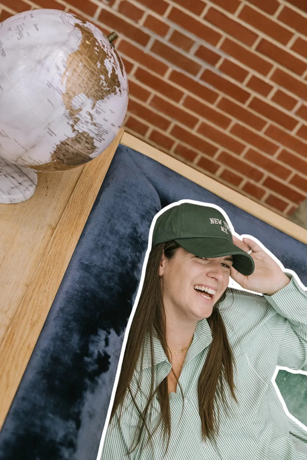 A woman with long brown hair, wearing a green cap with white text, a green and white striped shirt, and a gold necklace, is lying on a velvet blue cushion. She is smiling and holding her cap with her right hand. Near her on a wooden table, there is a globe with a marble base, against a red brick wall.