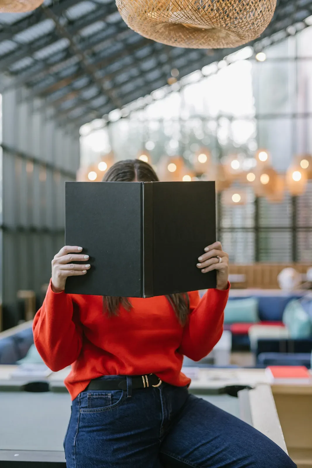 Person with long brown hair wearing a red long-sleeve shirt and dark jeans, sitting with one leg up, holding an open black book in front of their face in a modern, well-lit indoor space with wooden accents and hanging lights.