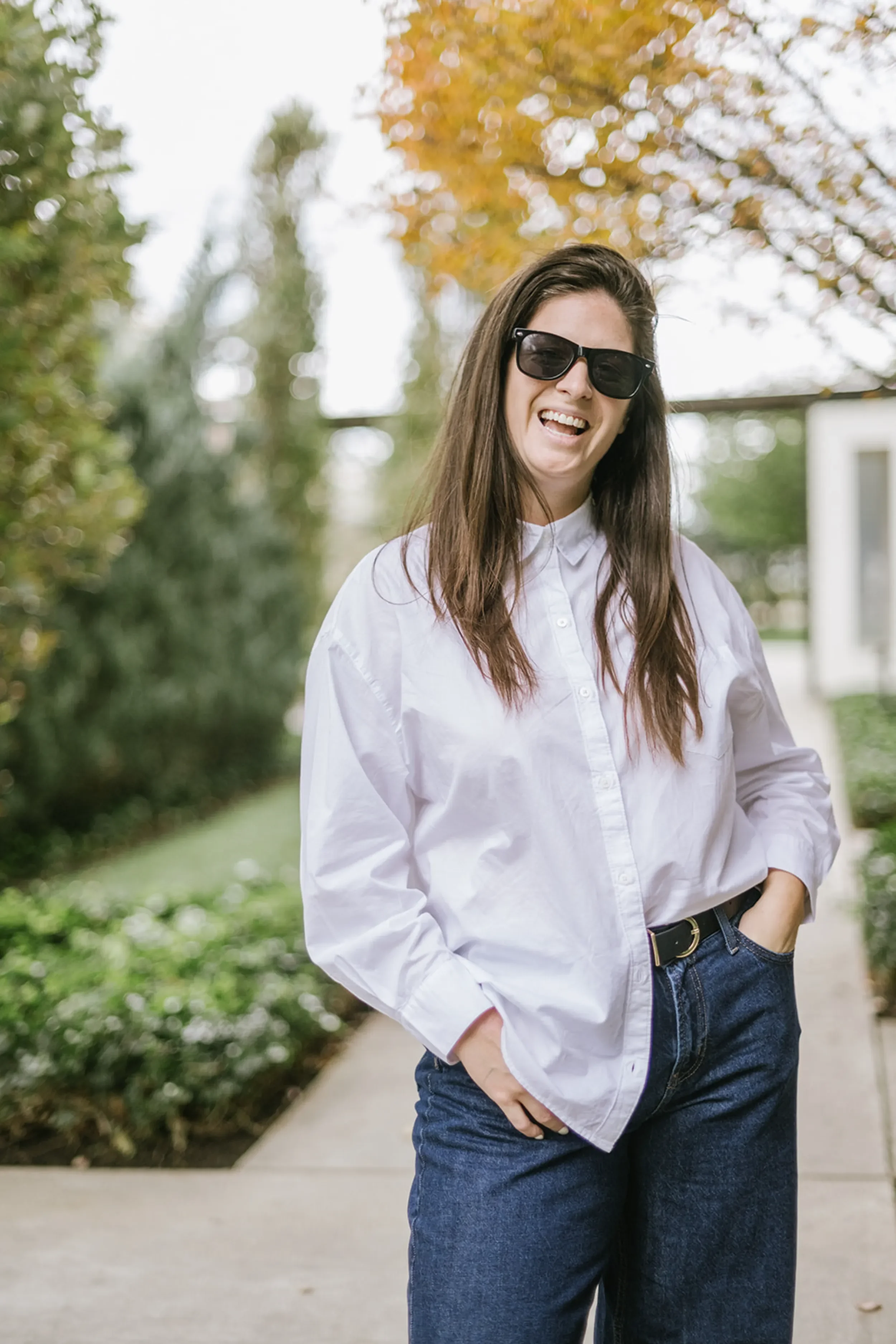 Woman with long brown hair, wearing sunglasses, a white button-up shirt, and blue jeans, standing outdoors on a sidewalk with trees in the background, smiling.