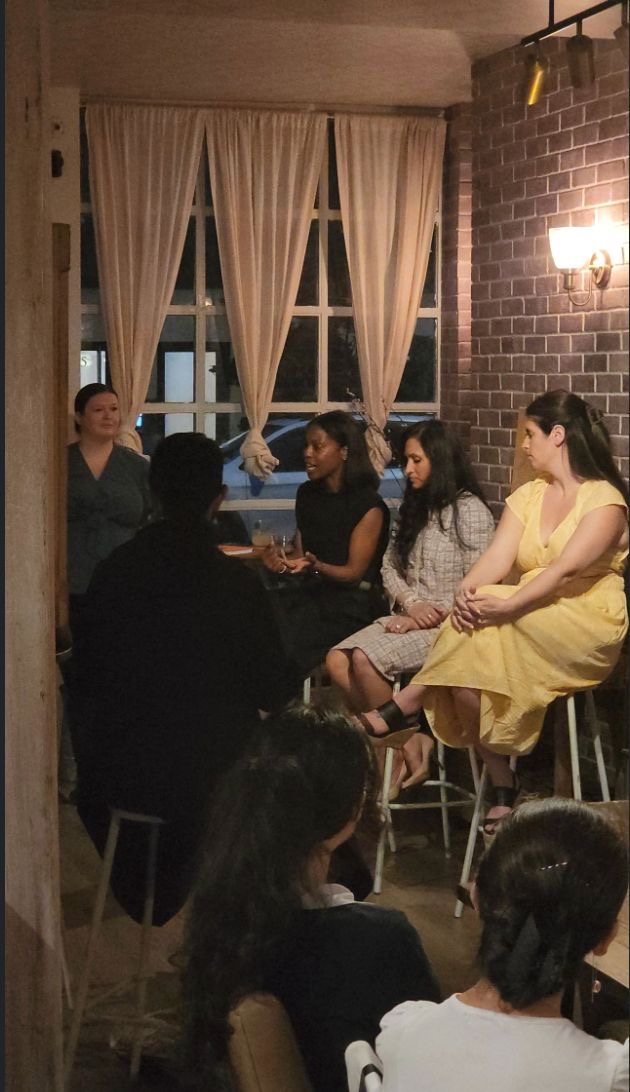 Group of women sitting and standing in a cozy indoor space with brick walls and large windows with curtains, engaged in conversation.