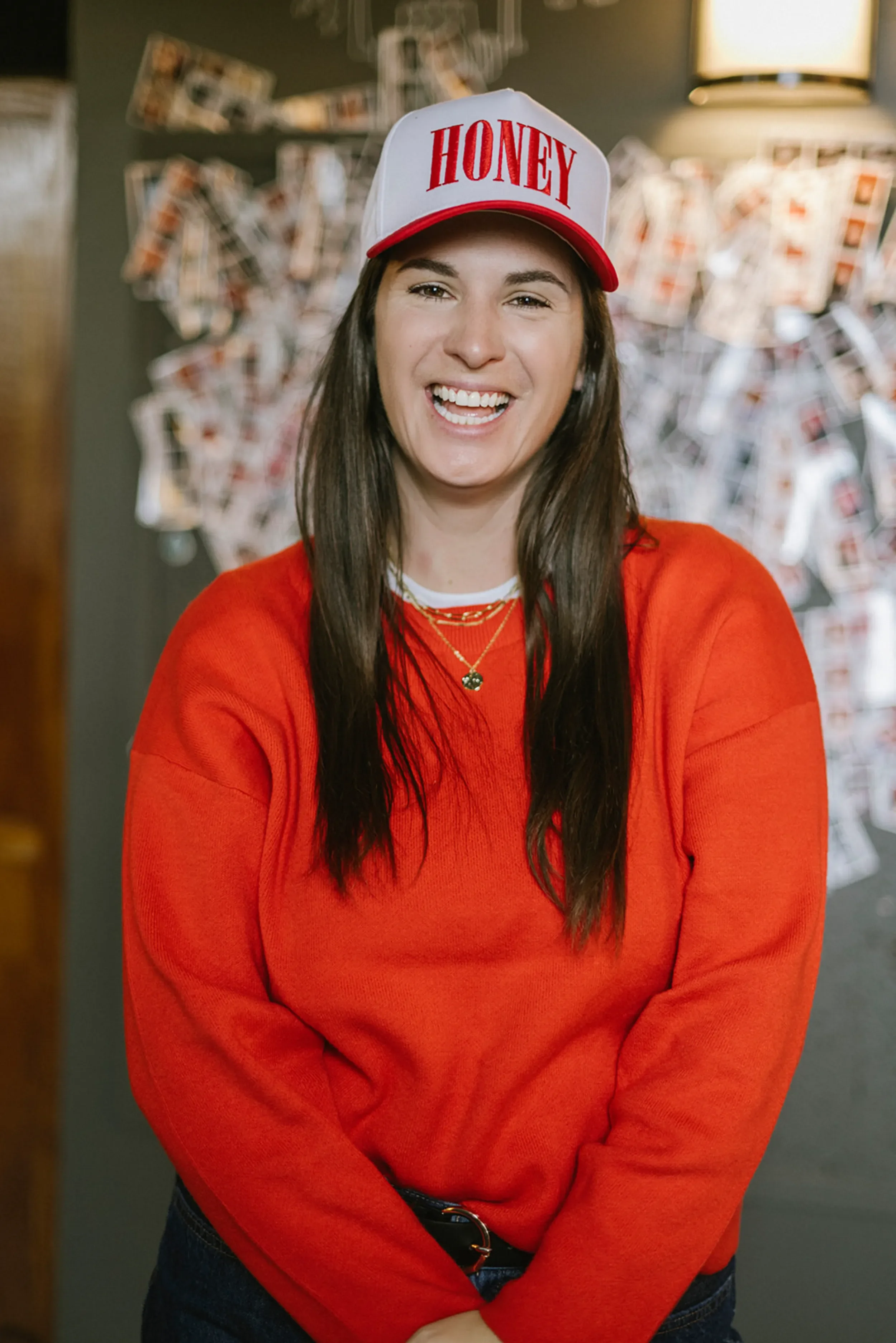 A woman with long brown hair wearing a white and red baseball cap that says "HONEY" in red letters, an orange sweater, and layered necklaces, smiling in front of a background with numerous photographs on a wall.