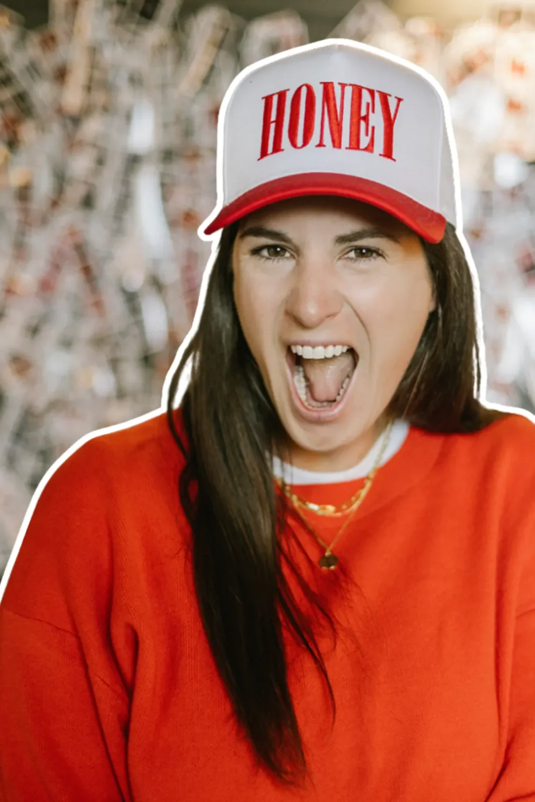 A woman wearing a red jacket and a white cap with red lettering that says 'HONEY', smiling and shouting with her mouth open, against a blurred outdoor background.
