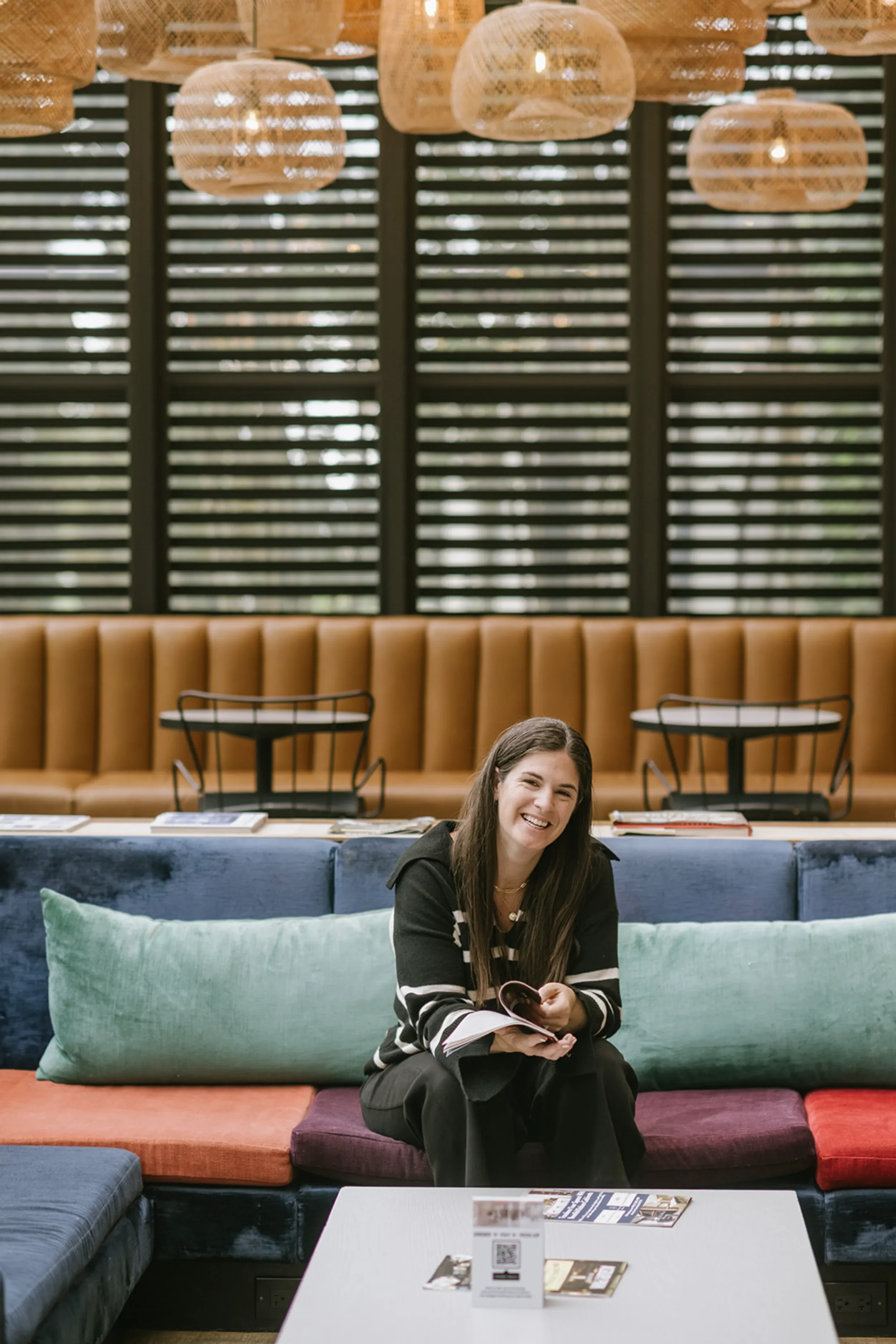 A woman sitting on a colorful sofa holding an open book, smiling at the camera, with a modern, stylish lounge background including a wooden slat wall, hanging lights, and a high cushioned backrest.
