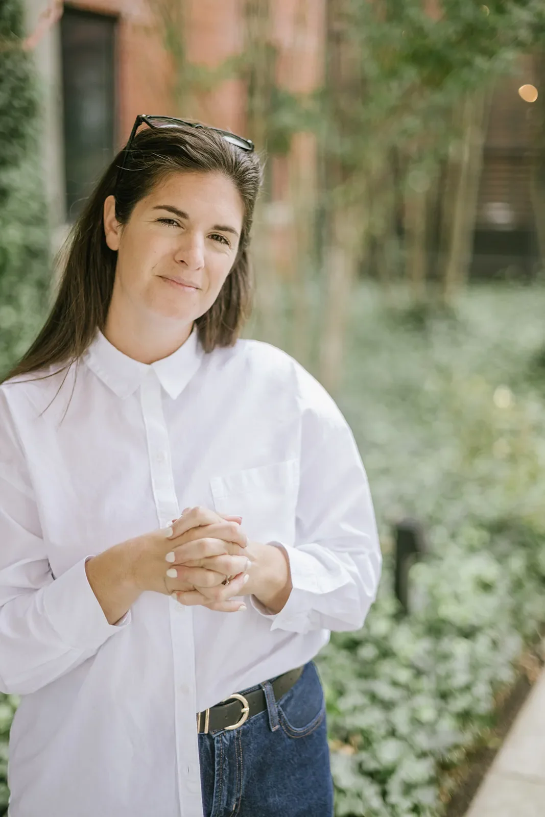 A woman with sunglasses on her head wearing a white button-up shirt and jeans, standing outdoors with greenery in the background.