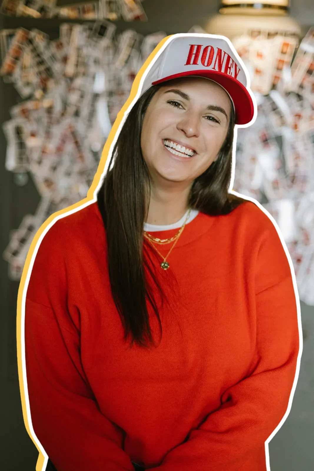 Young woman smiling, wearing a red sweatshirt and a white and red hat with 'HONEY' written on it, standing in front of a wall with hanging photographs or postcards.