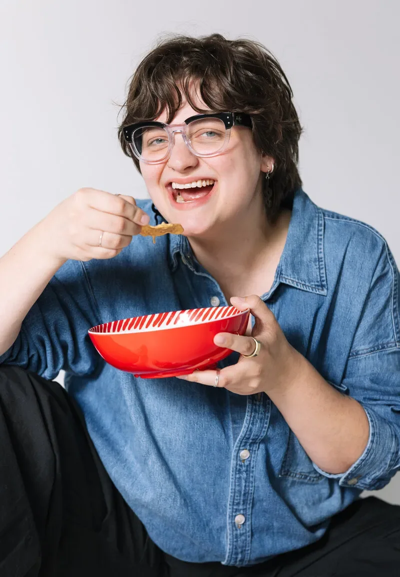 Person with short curly hair and glasses wearing a blue denim shirt, smiling and holding a red bowl with food while eating.