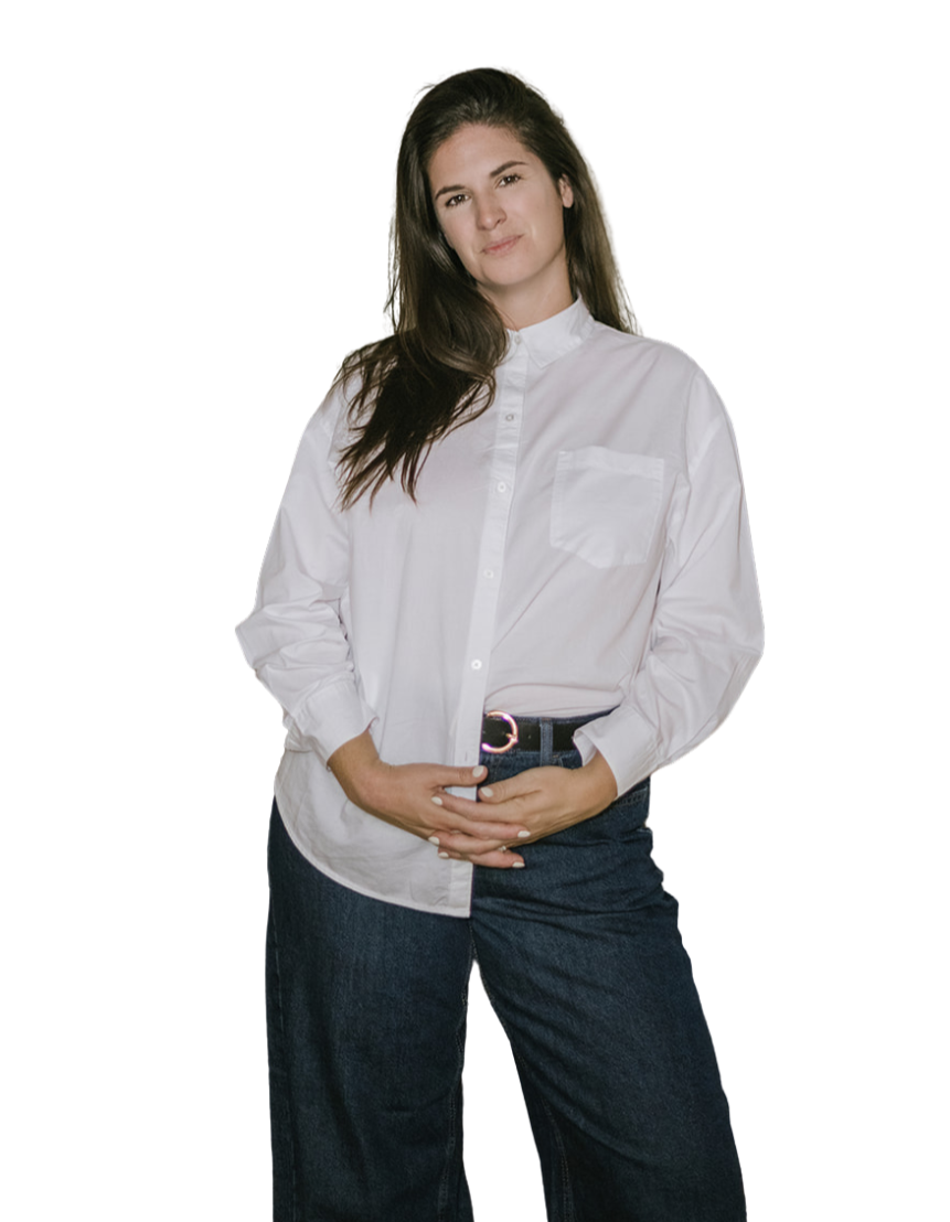 Young woman with long brown hair wearing a white button-up shirt and dark jeans, standing with hands clasped in front, looking confident.
