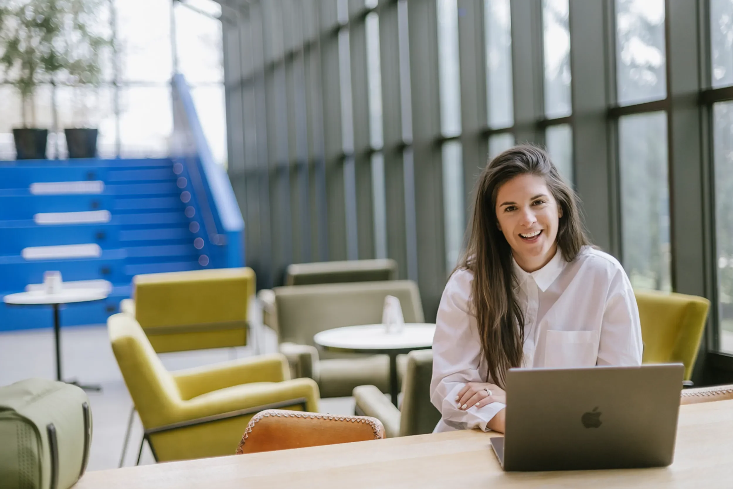 A young woman with long dark hair sitting at a table with a MacBook laptop, smiling in a modern, brightly lit indoor space with large windows and colorful armchairs.