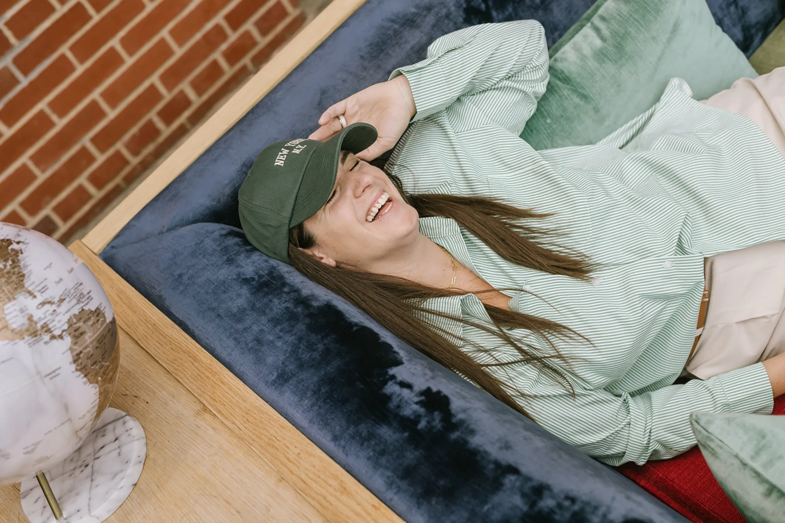 A woman lying on a sofa, smiling and laughing, wearing a green cap and striped shirt, with a globe on the wooden table nearby.