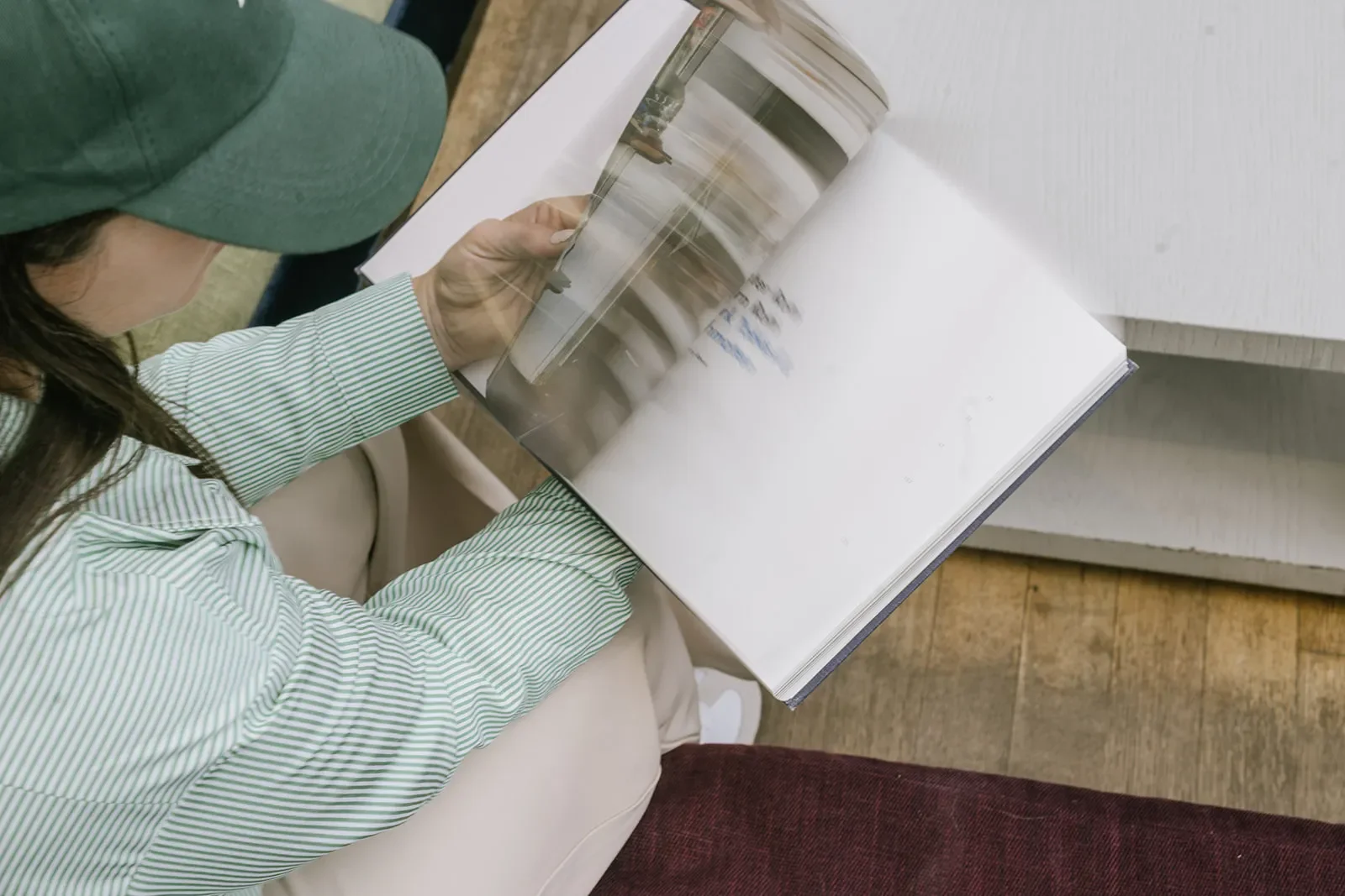 Person wearing a green cap and green striped shirt sitting on a chair, looking at an open photo album with photographs on white sheet pages.