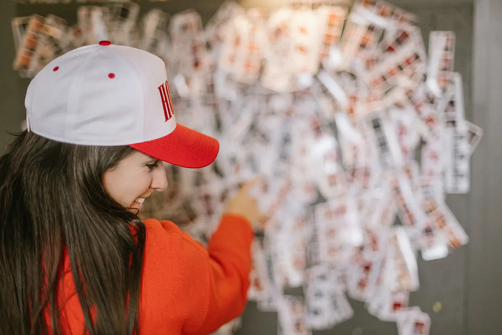 A girl smiling and pointing at a large display of scattered playing cards pinned to a gray wall, wearing a white and red cap and red jacket.