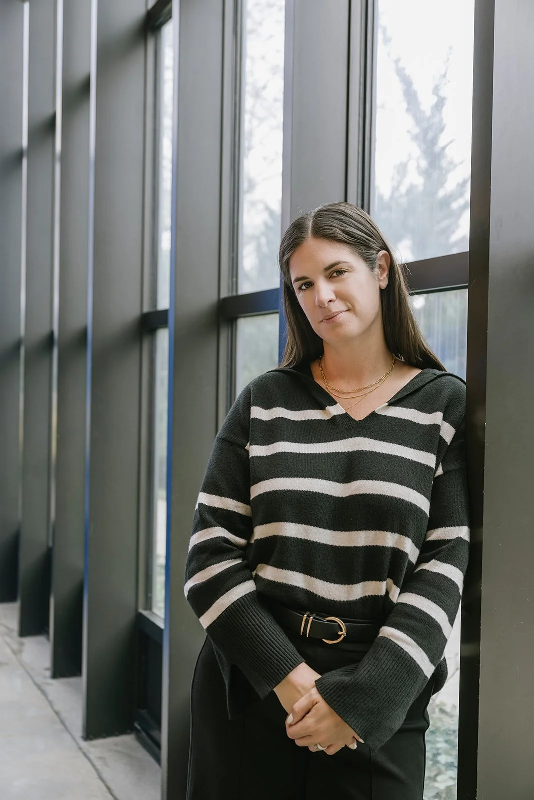A woman with long brown hair wearing a black and white striped sweater and black pants, standing indoors against large windows.