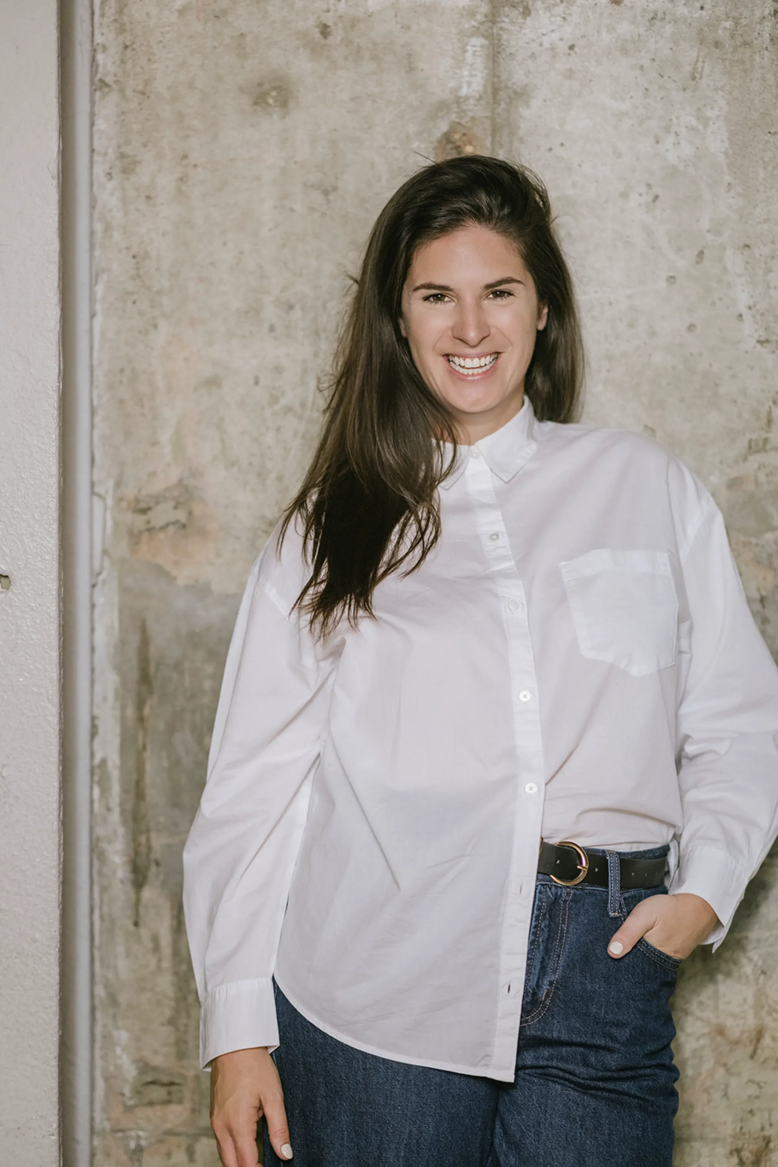 A woman with long brown hair smiling and wearing a white button-up shirt and jeans standing against a textured wall.