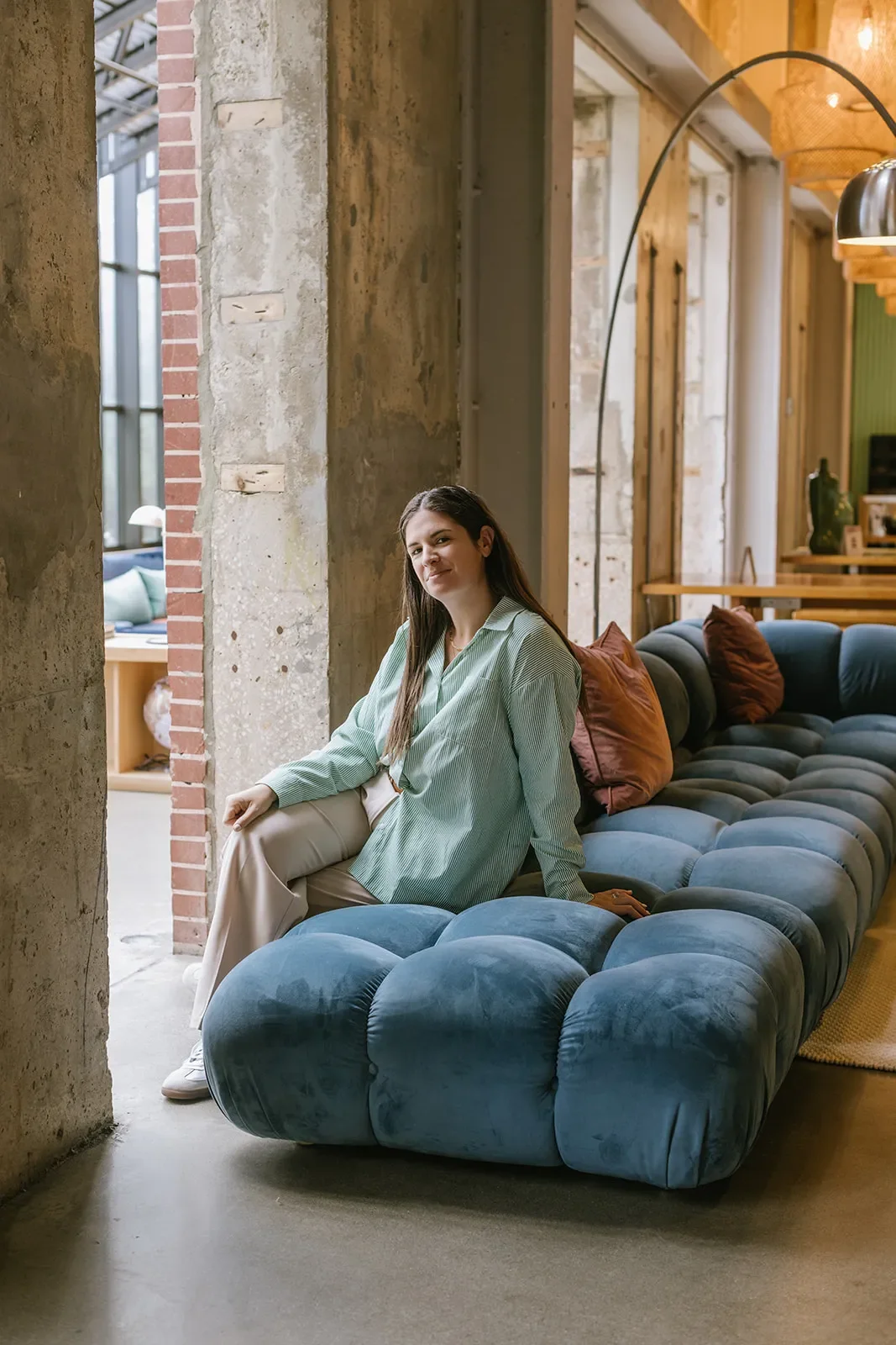 A woman with long brown hair sitting on a blue velvet tufted sofa in a modern industrial-style room. She is wearing a light green striped shirt and beige pants, smiling and looking at the camera, with a large window and brick and concrete walls behind her.