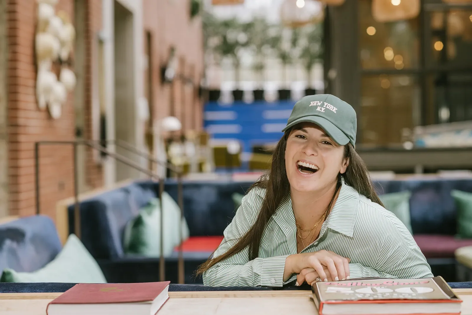 A young woman with long dark hair, smiling and laughing, sitting at a table inside a cafe. She is wearing a gray baseball cap that says 'New York N.Y.' and a striped shirt. In front of her are two books, and the background shows a cozy cafe interior with brick walls, cushions, and a large window.