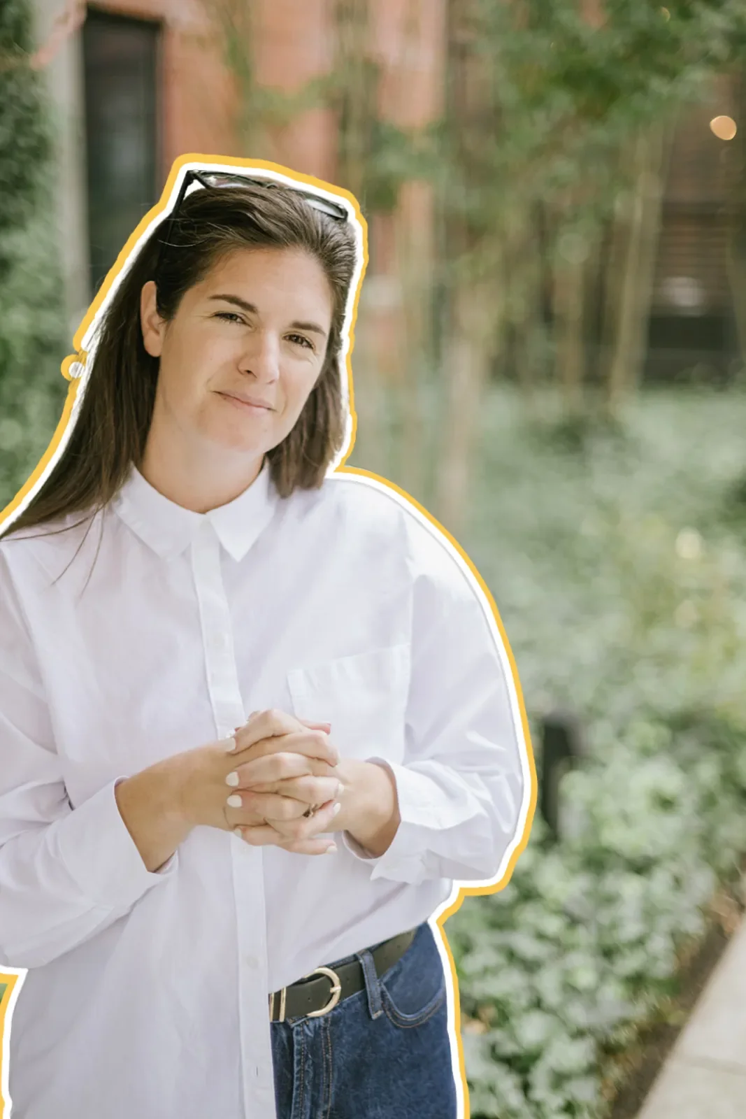 A woman with long brown hair, white shirt, and black belt, standing outdoors with greenery and a building in the background, looking at the camera with a slight smile.