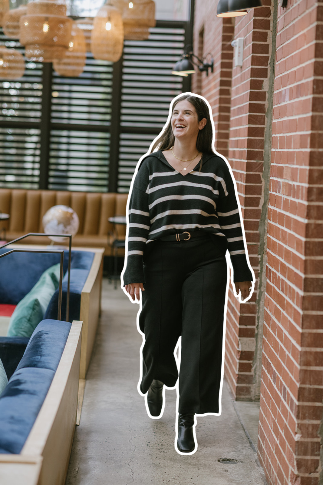 A woman with long dark hair and a big smile wearing a black and white striped sweater, black pants, and black boots walking near a brick wall in a stylish indoor space.
