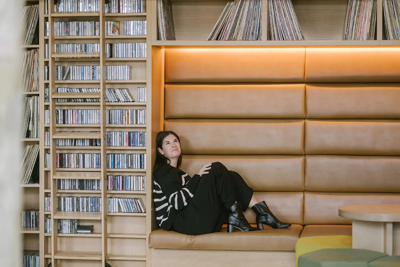 A woman sitting on a leather bench against a padded wall in a modern interior space, surrounded by wooden bookshelves filled with CDs and vinyl records.