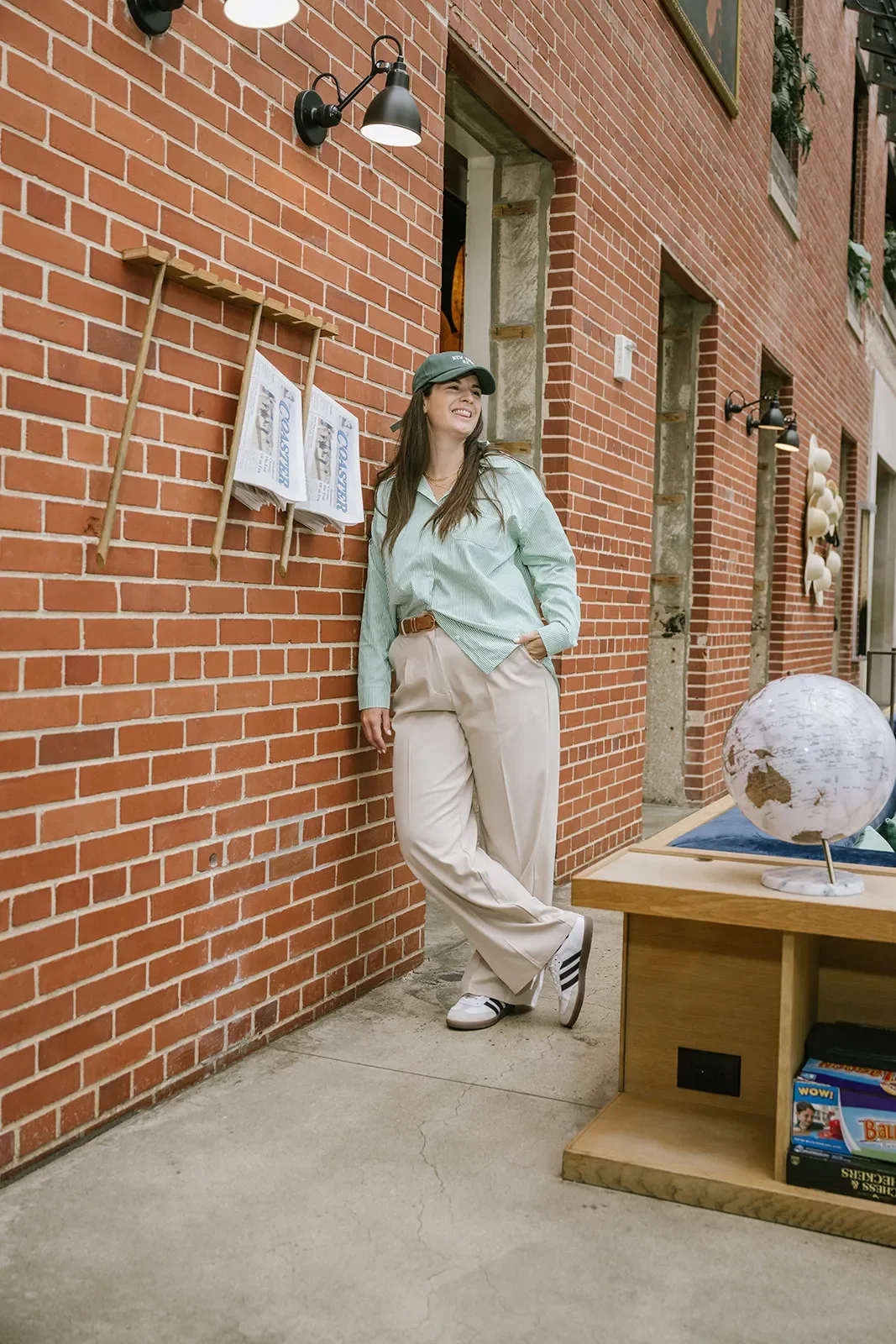 A woman leaning against a brick wall, smiling, wearing a green cap, light green shirt, beige pants, and sneakers, with newspapers on a wooden rack behind her and a globe on a table nearby.