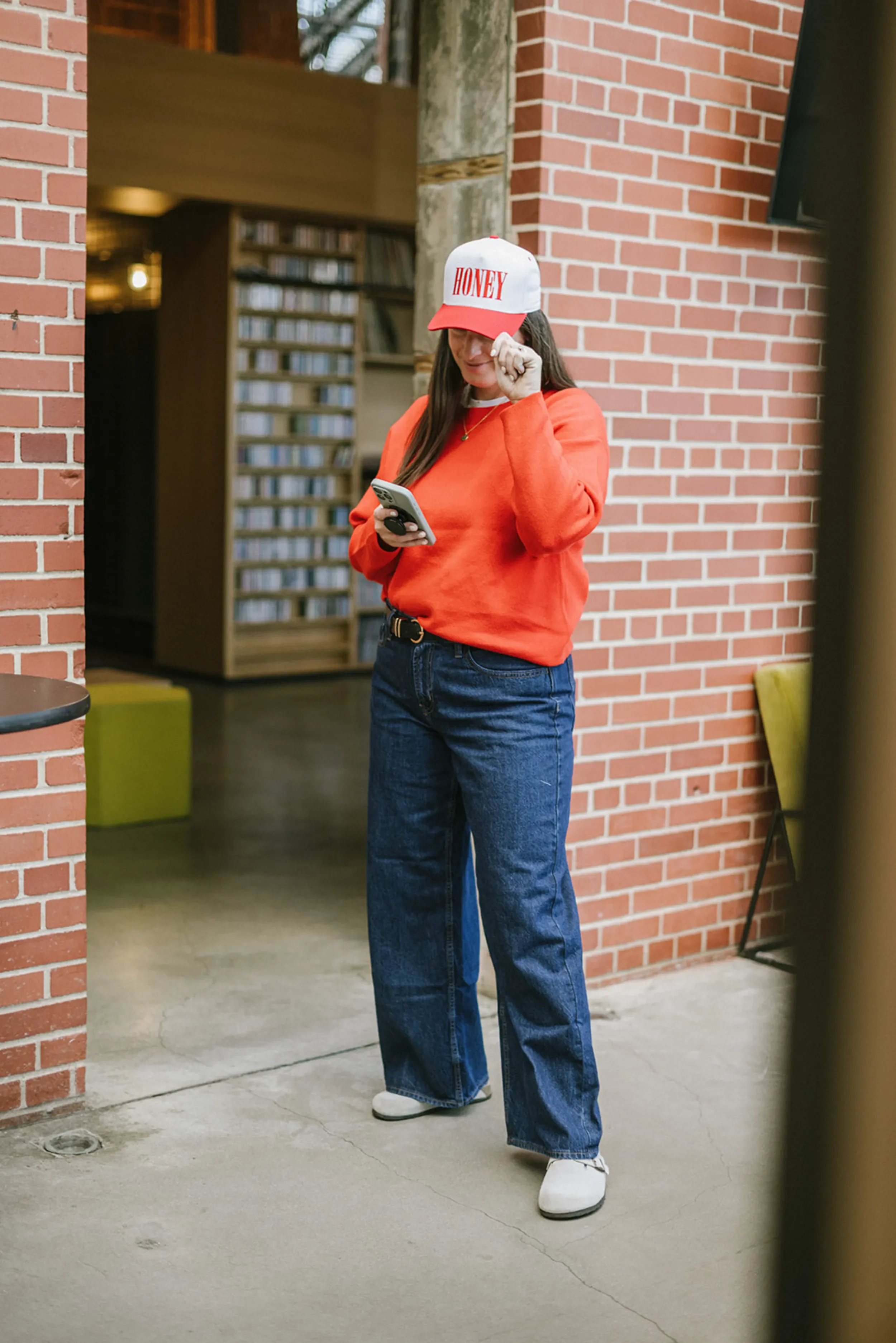 Woman wearing a white and red cap labeled 'HONEY,' orange sweatshirt, blue jeans, and white shoes, standing inside near a brick wall and looking at her phone.