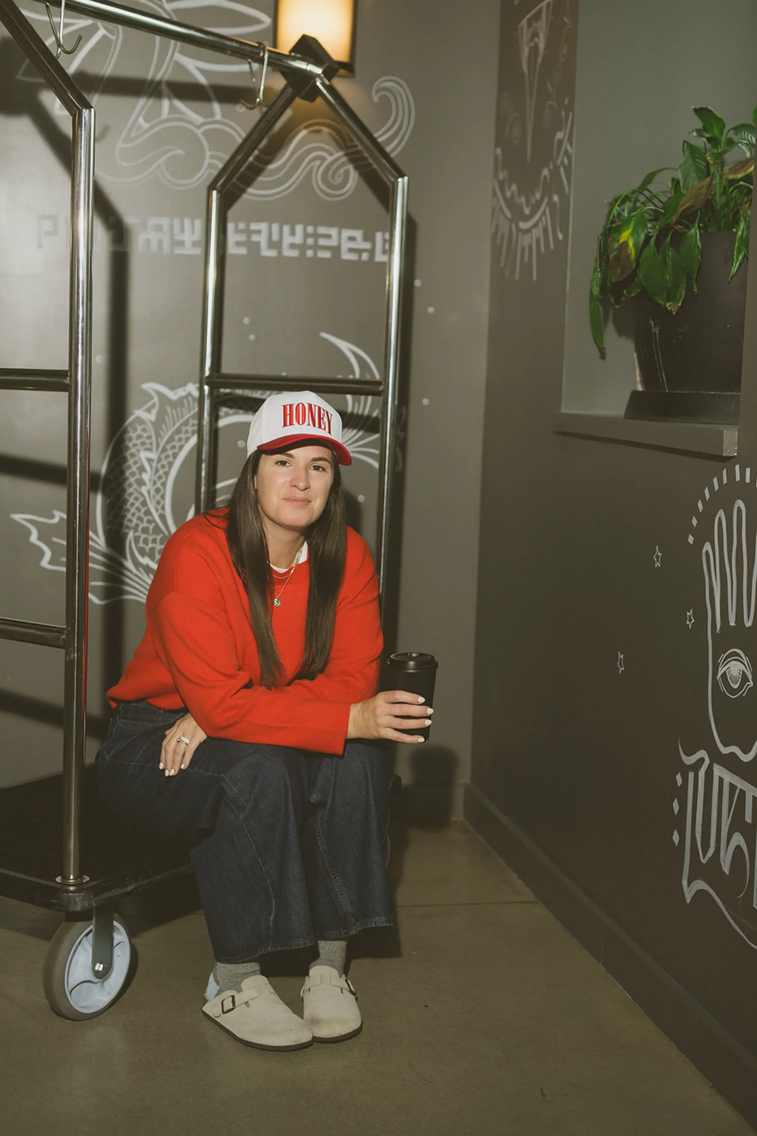 A woman wearing a white cap with red lettering, a red sweater, and dark jeans sitting on a luggage cart in a cafe or restaurant. She is holding a black coffee cup, surrounded by dark walls with white chalk art and a potted plant.