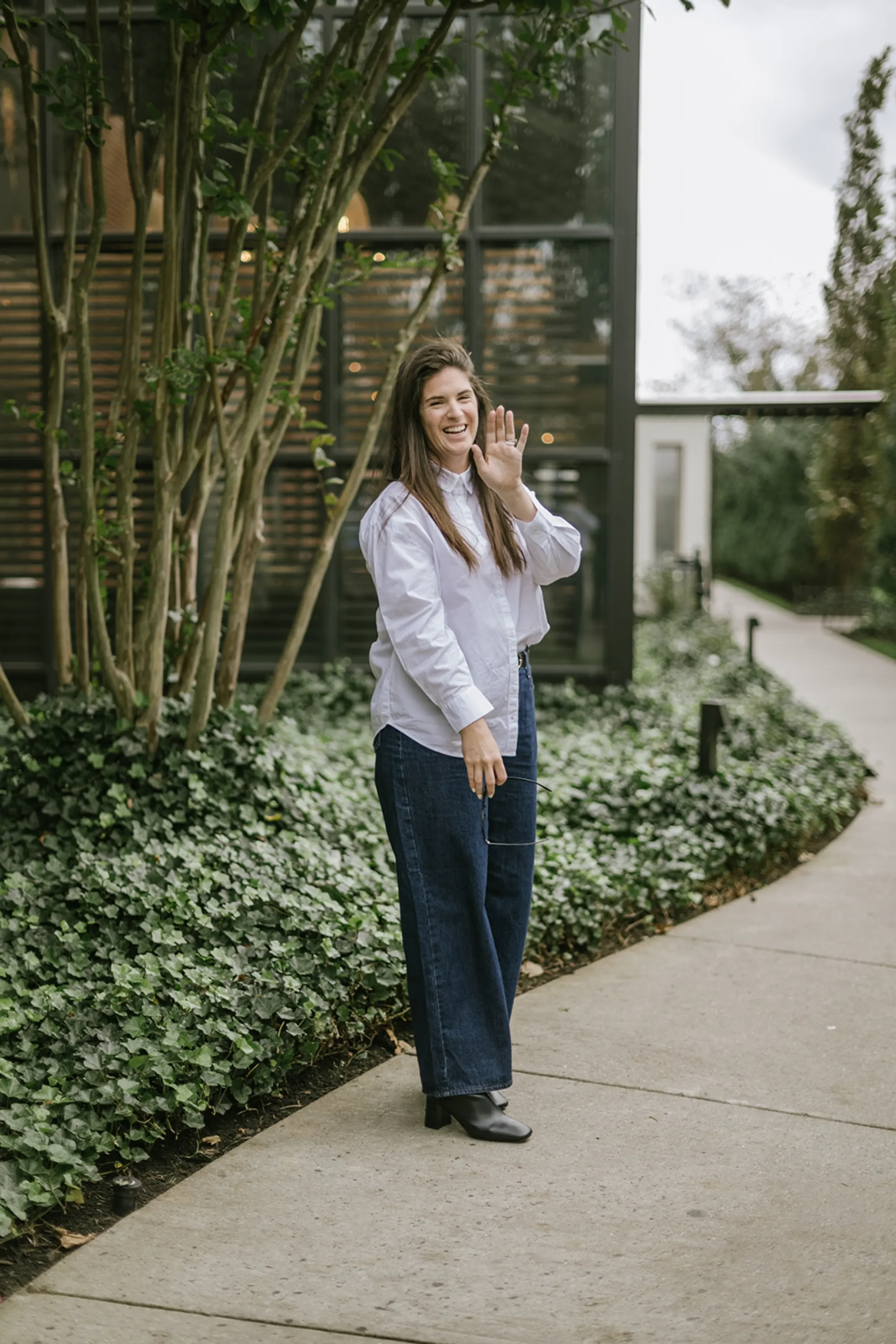 A woman with long brown hair wearing a white blouse, dark jeans, and black heeled boots, standing on a sidewalk beside green plants and trees, smiling and waving.