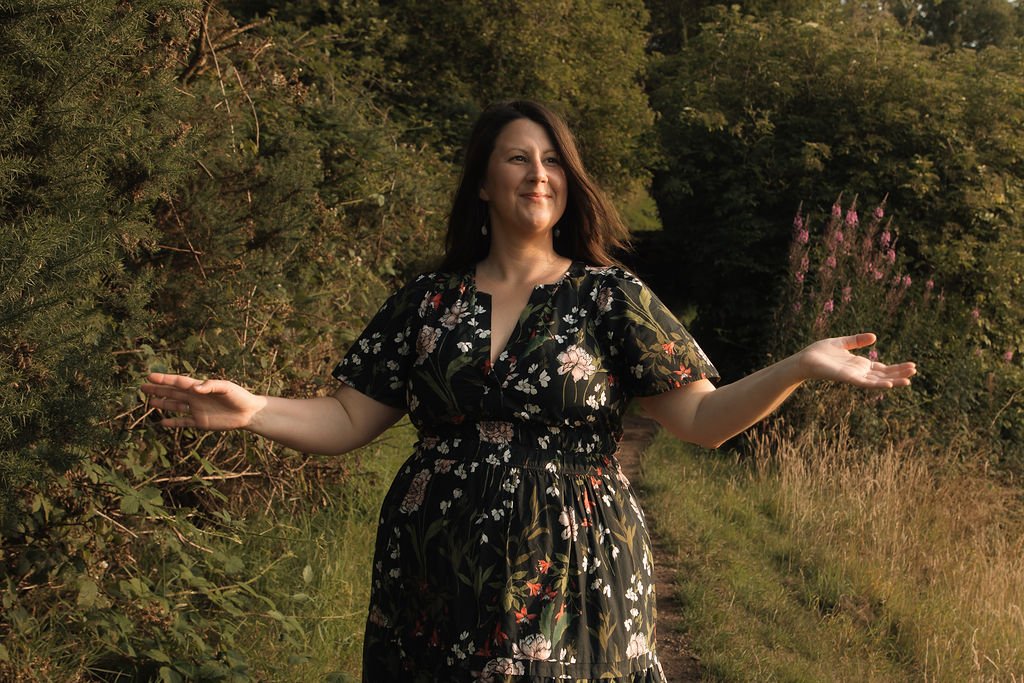 A woman in a black floral dress standing on a grassy path with her arms outstretched, smiling, surrounded by green trees and plants in a natural outdoor setting.