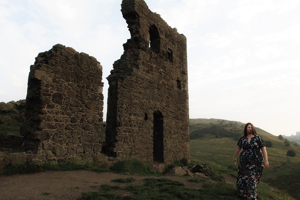 A woman walking on a grassy hill with an ancient stone ruin in the background.