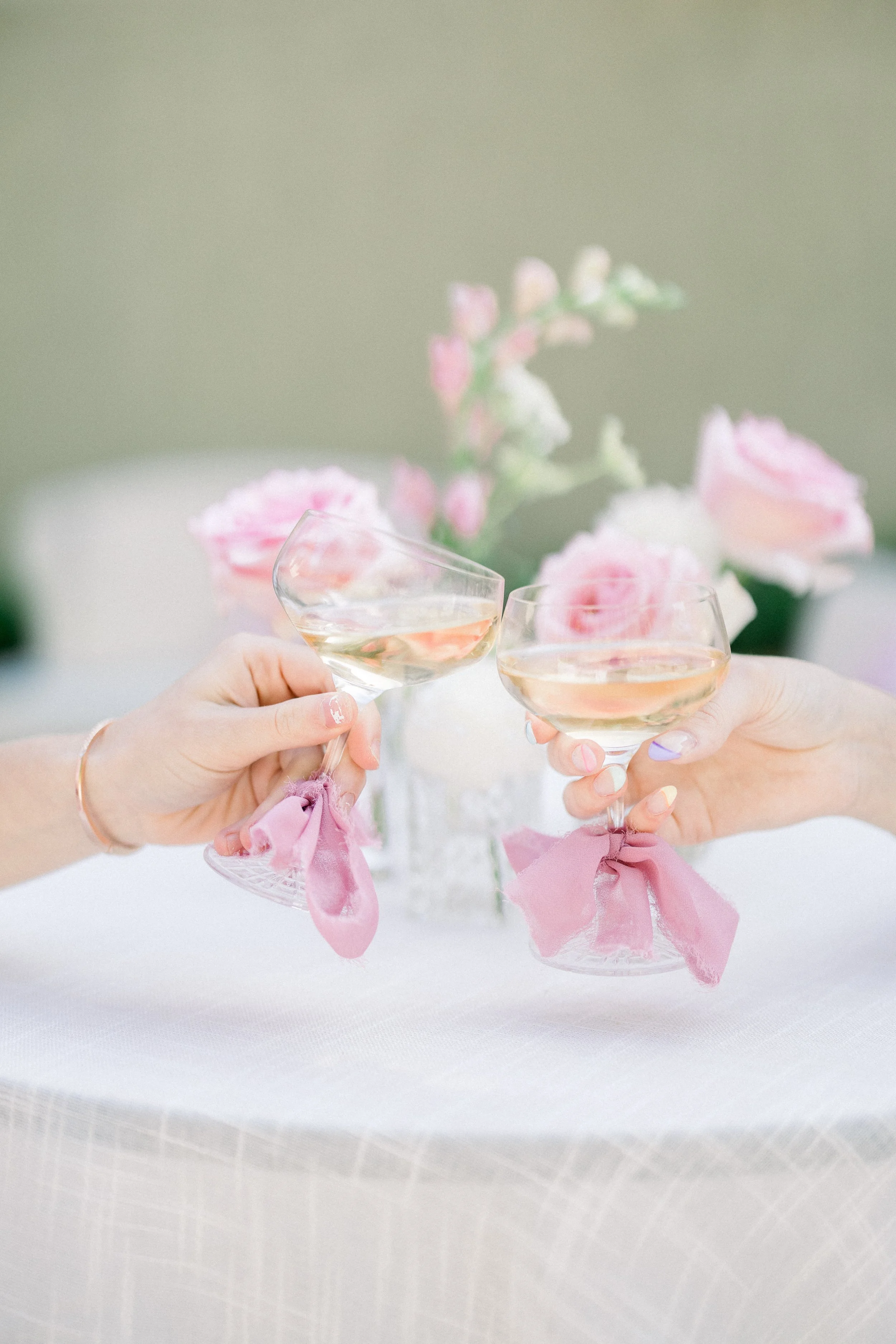 Two hands holding champagne glasses with pink ribbons, toasting over a white table with pink flowers in the background.