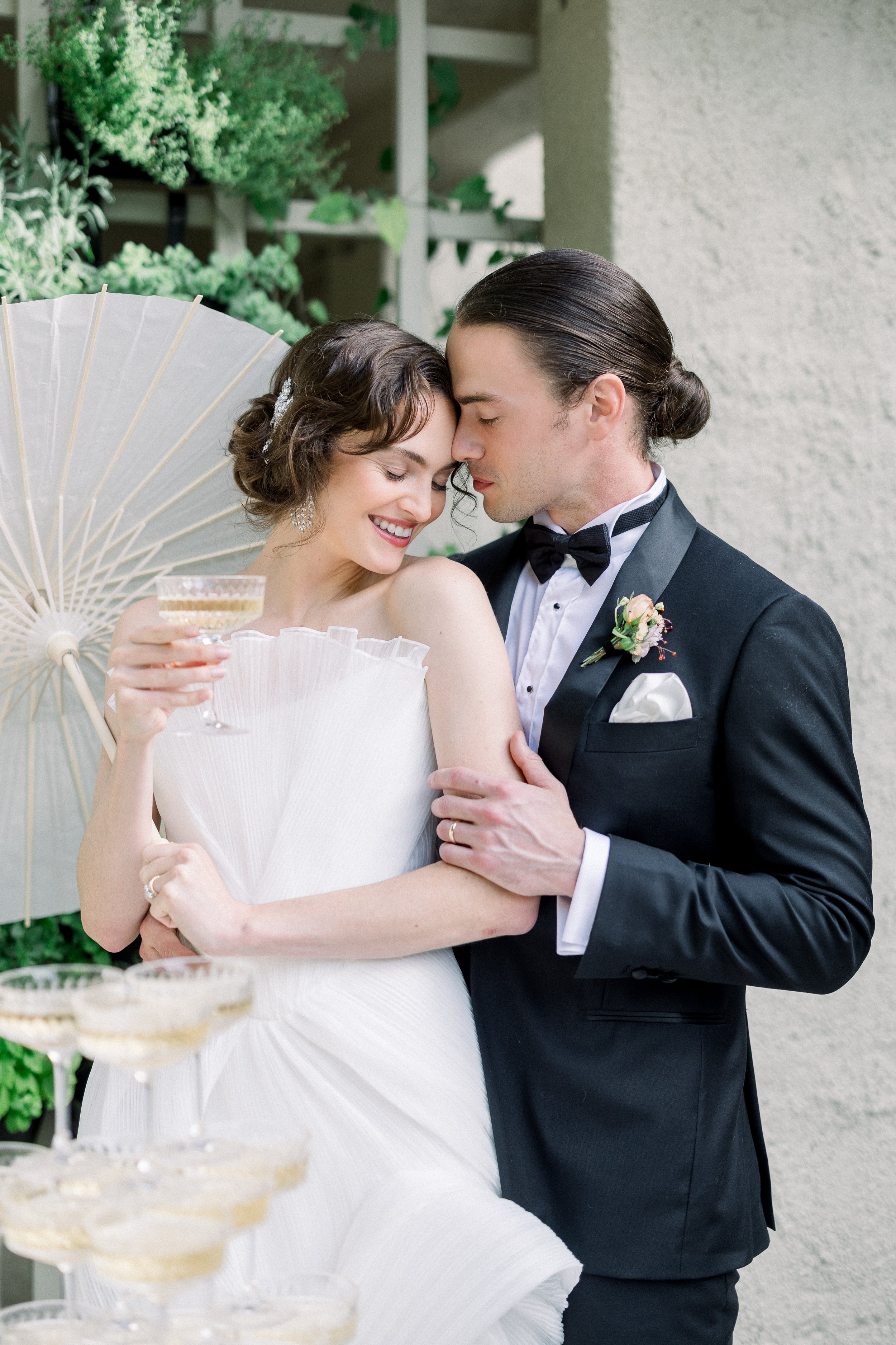 Bride and groom embracing, she holds a champagne glass, near a champagne tower and parasol.