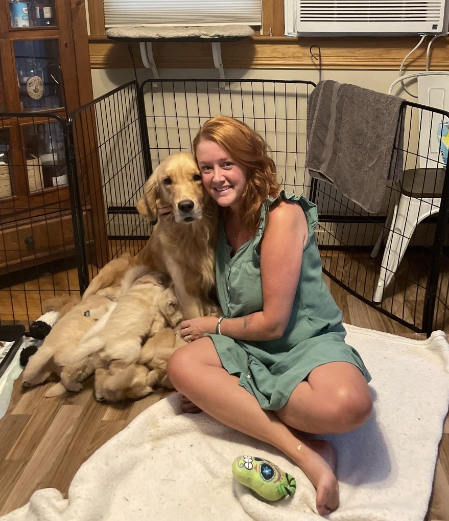 A woman with red hair sitting on the floor, smiling, among a litter of golden retriever puppies, inside a playpen in a home.