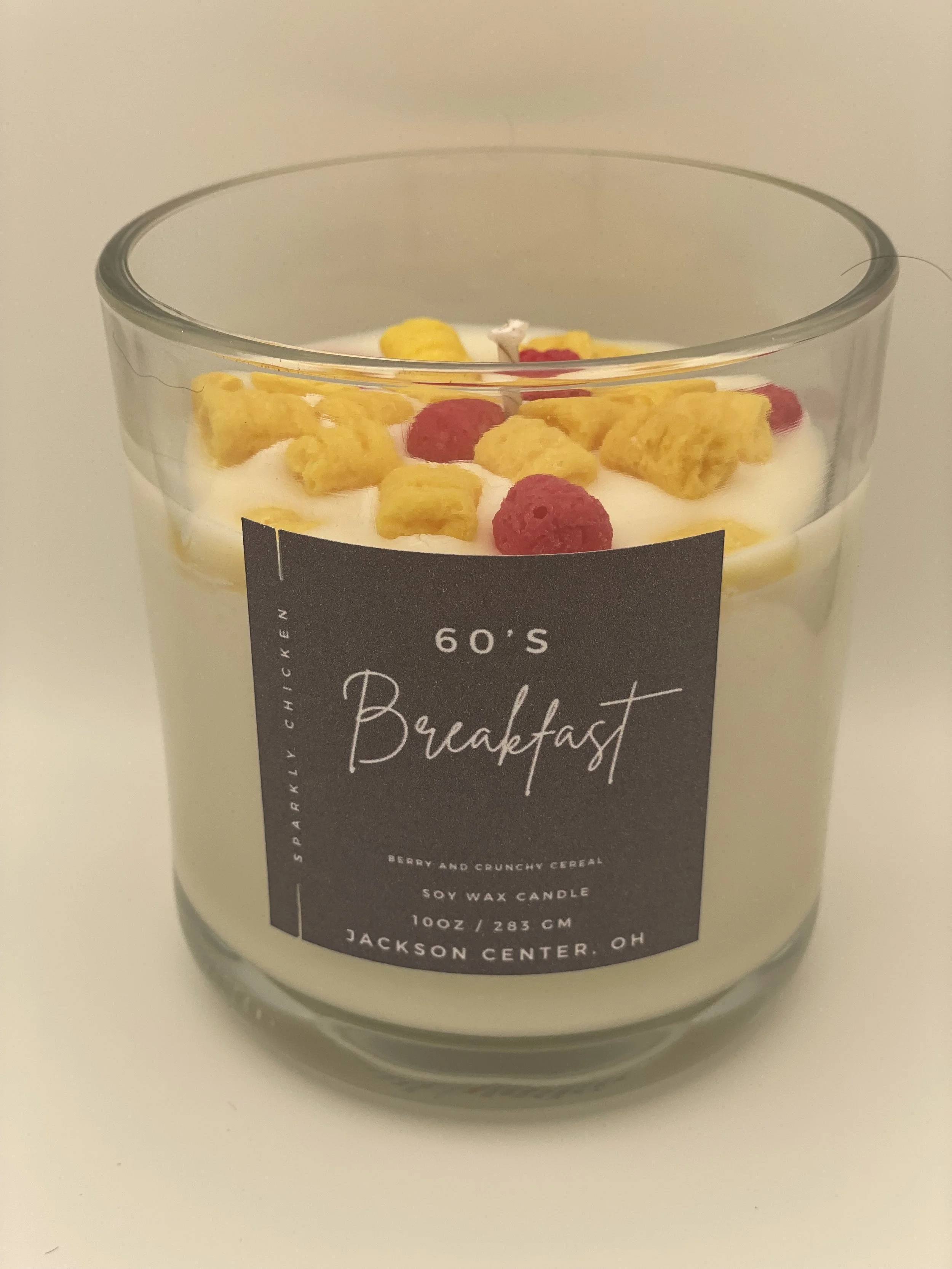 A scented candle in a glass jar labeled '60's Breakfast,' topped with dried cereal pieces and raspberries.