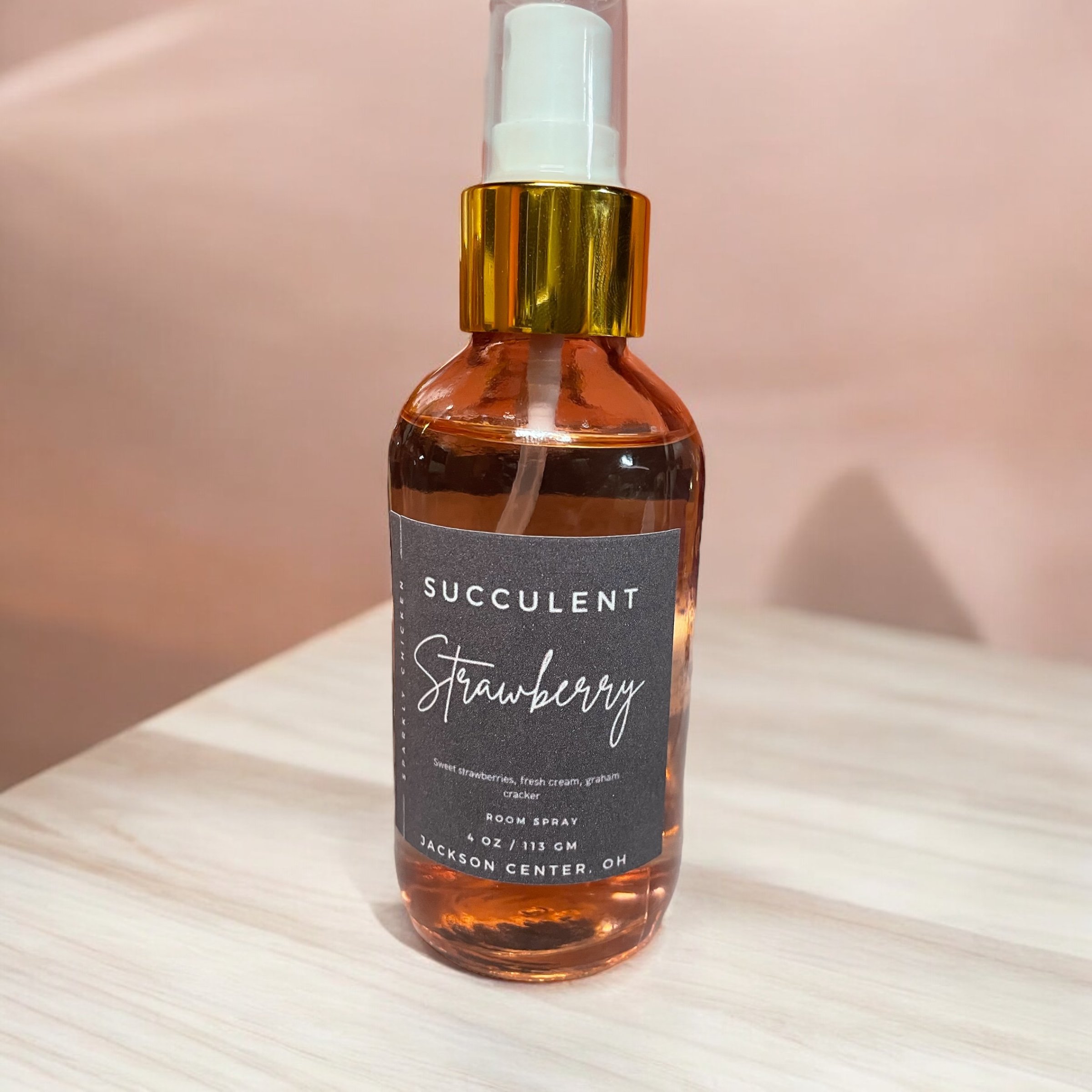 A clear plastic spray bottle with pink liquid inside and a gold and white spray nozzle. The label reads 'Succulent Strawberry' and describes it as a room spray with a scent of strawberries, fresh cream, graham cracker. The bottle contains 4 oz. and is from Jackson Center, Ohio.