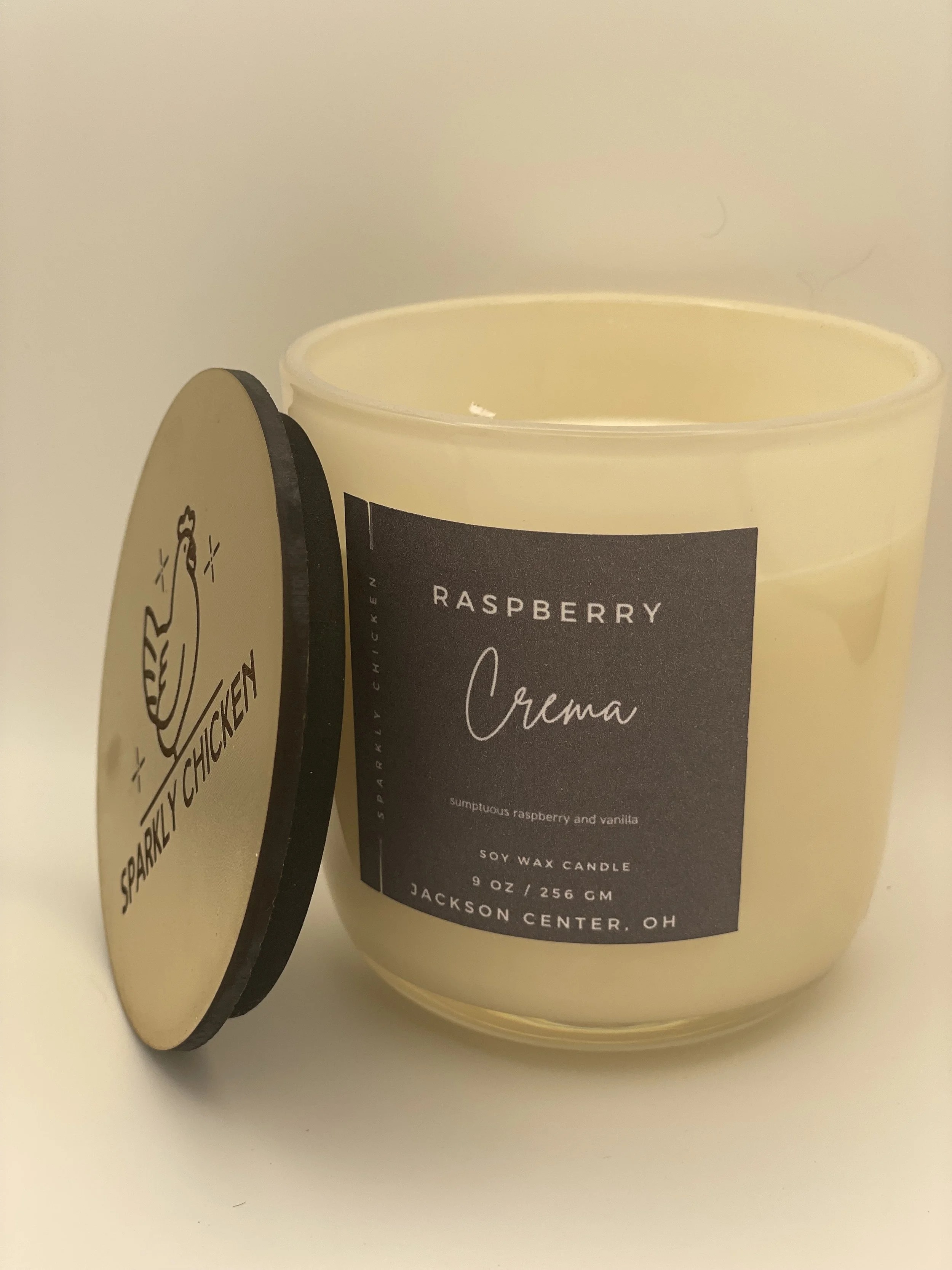 Cream-colored soy wax candle in a glass jar with a black label that reads 'Raspberry Crema' and 'scented with raspberry and vanilla.' The candle is from Sparkly Chicken, Jackson Center, Ohio, with a black lid beside it featuring a chicken logo and the brand name.