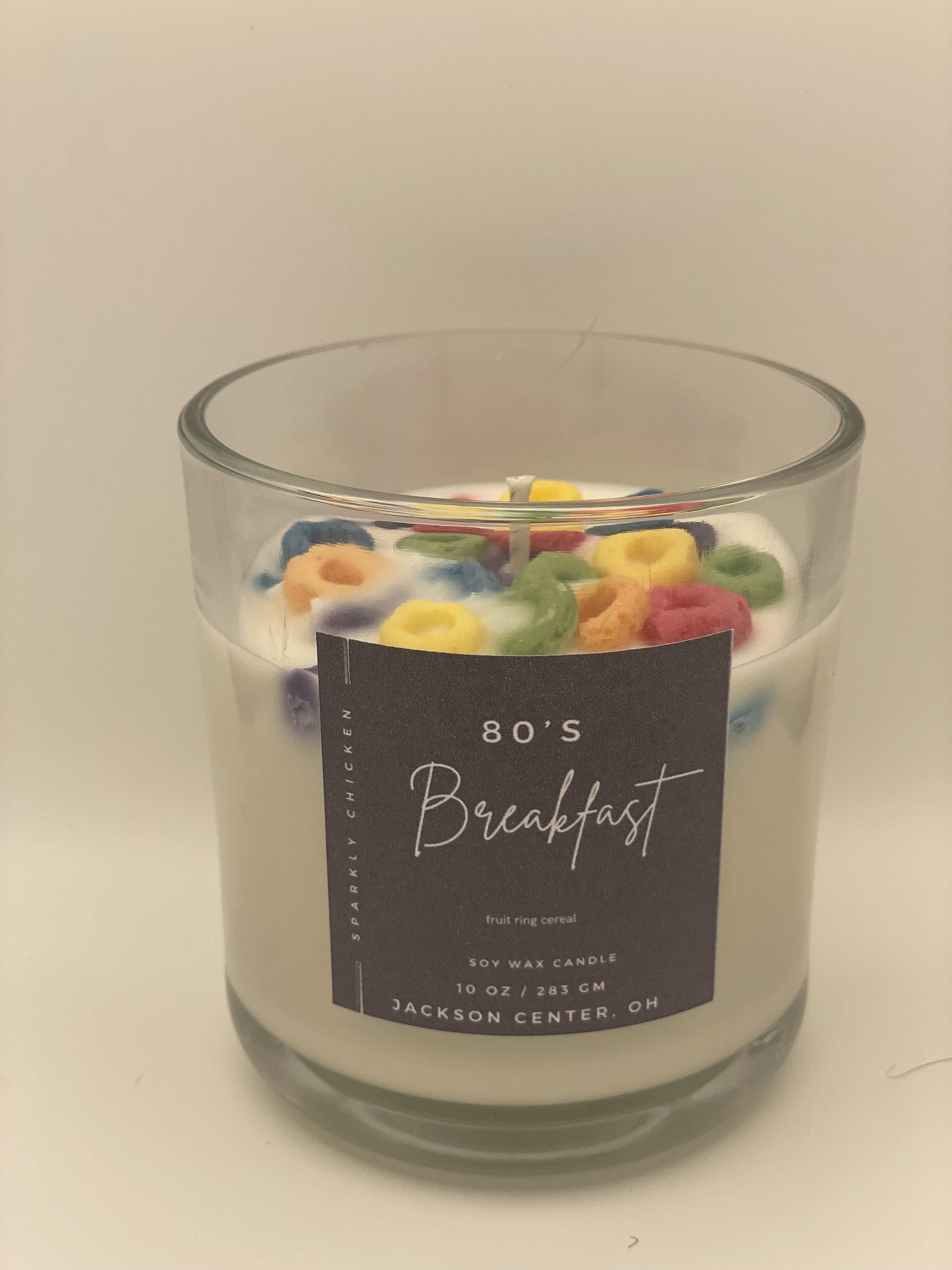 A glass candle with colorful fruit-shaped cereal pieces on top, labeled as '80's Breakfast, soy wax, 10 oz, Jackson Center, OH.'