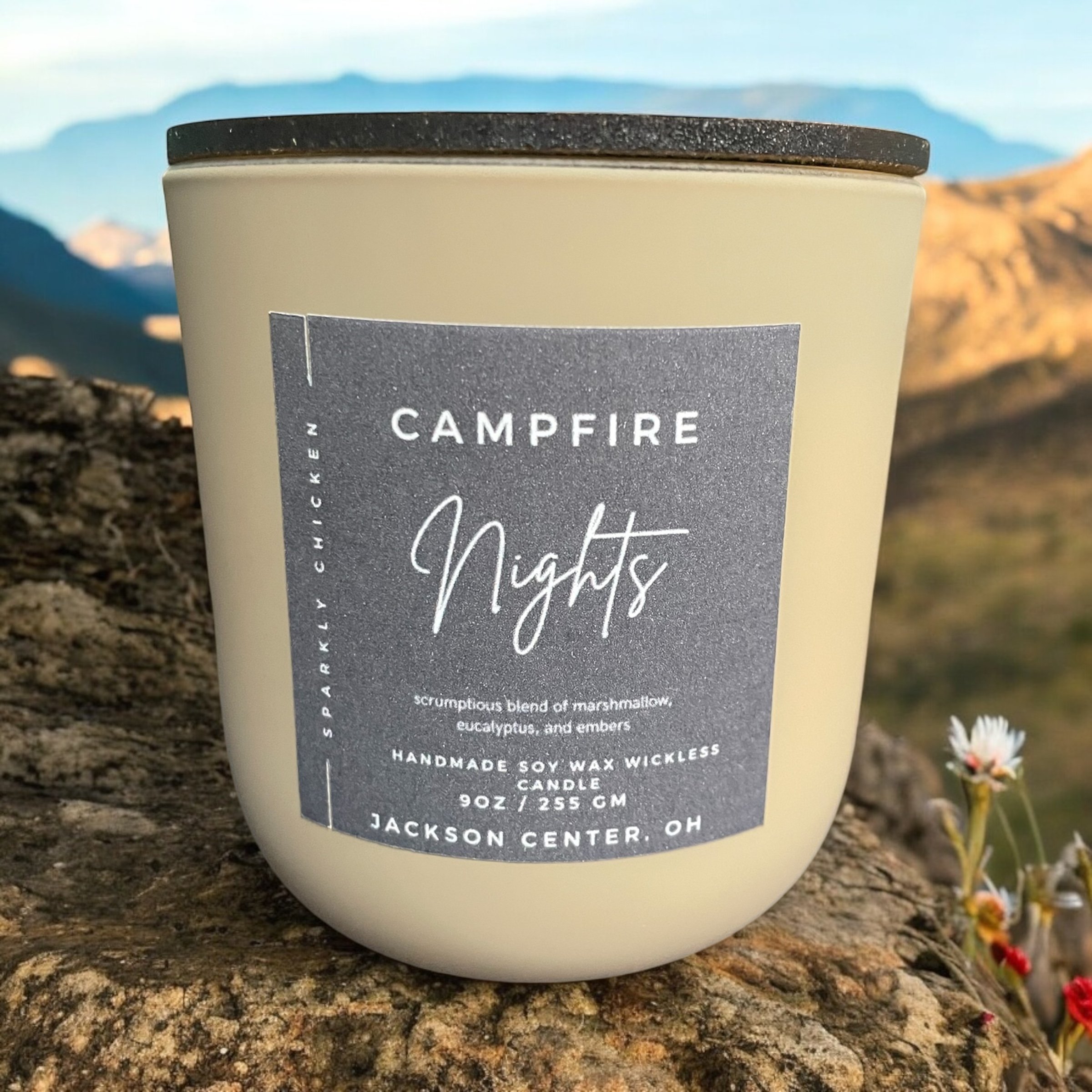 A cream-colored candle with a label reading "Campfire Nights" placed on a rock outdoors, with mountains and flowers in the background.