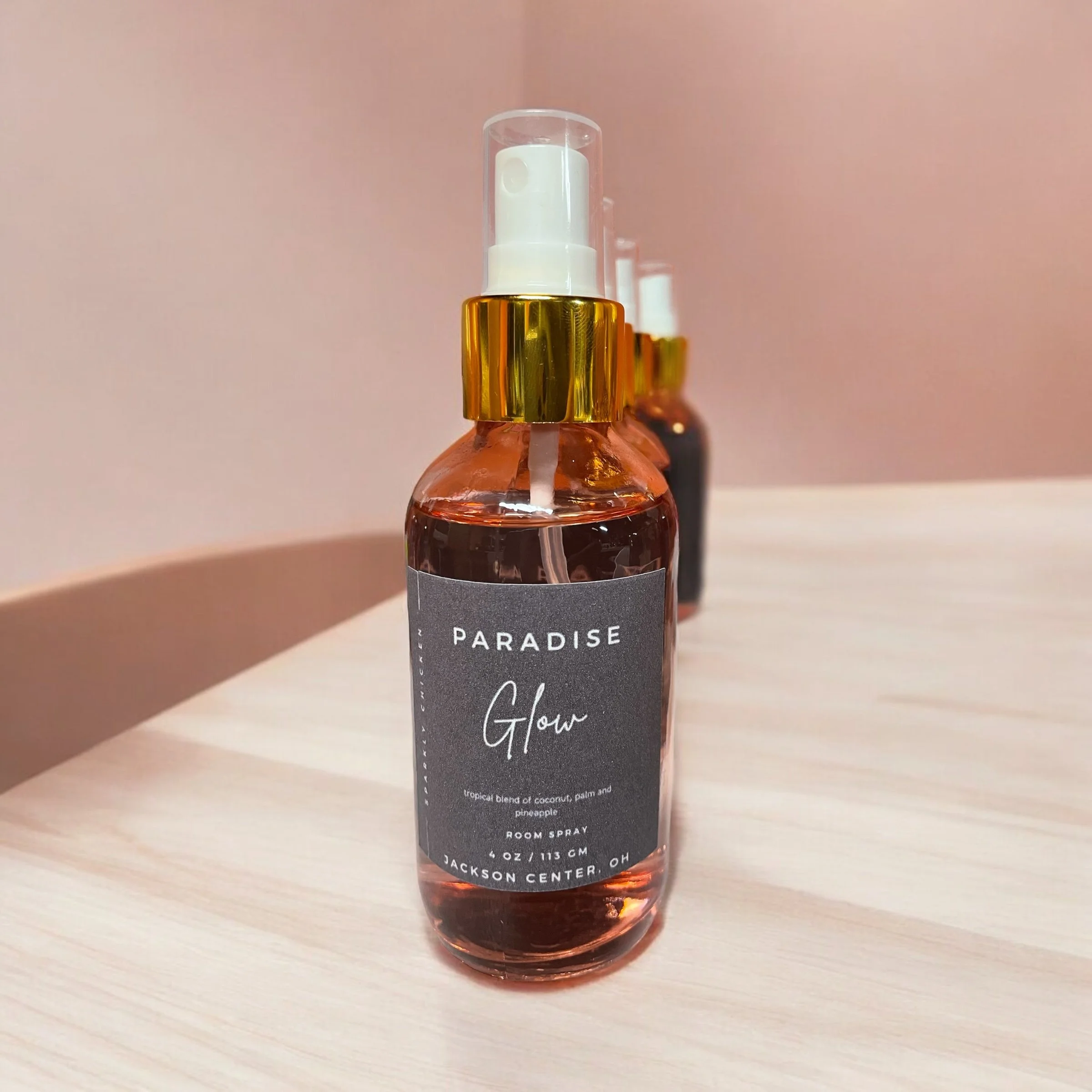 A spray bottle labeled 'Paradise Glow,' containing tropical blend room spray with coconut, palm, and pineapple scents, on a light wooden surface with two similar bottles in the background.
