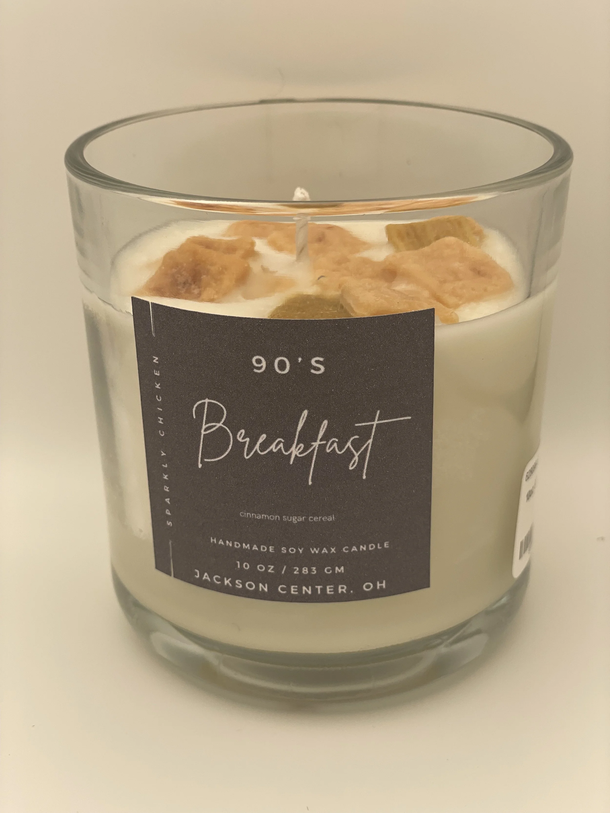 A scented candle in a glass jar with a black label, featuring the words '90's Breakfast' and description of cinnamon sugar cereal. The candle has decorative cereal pieces on top.