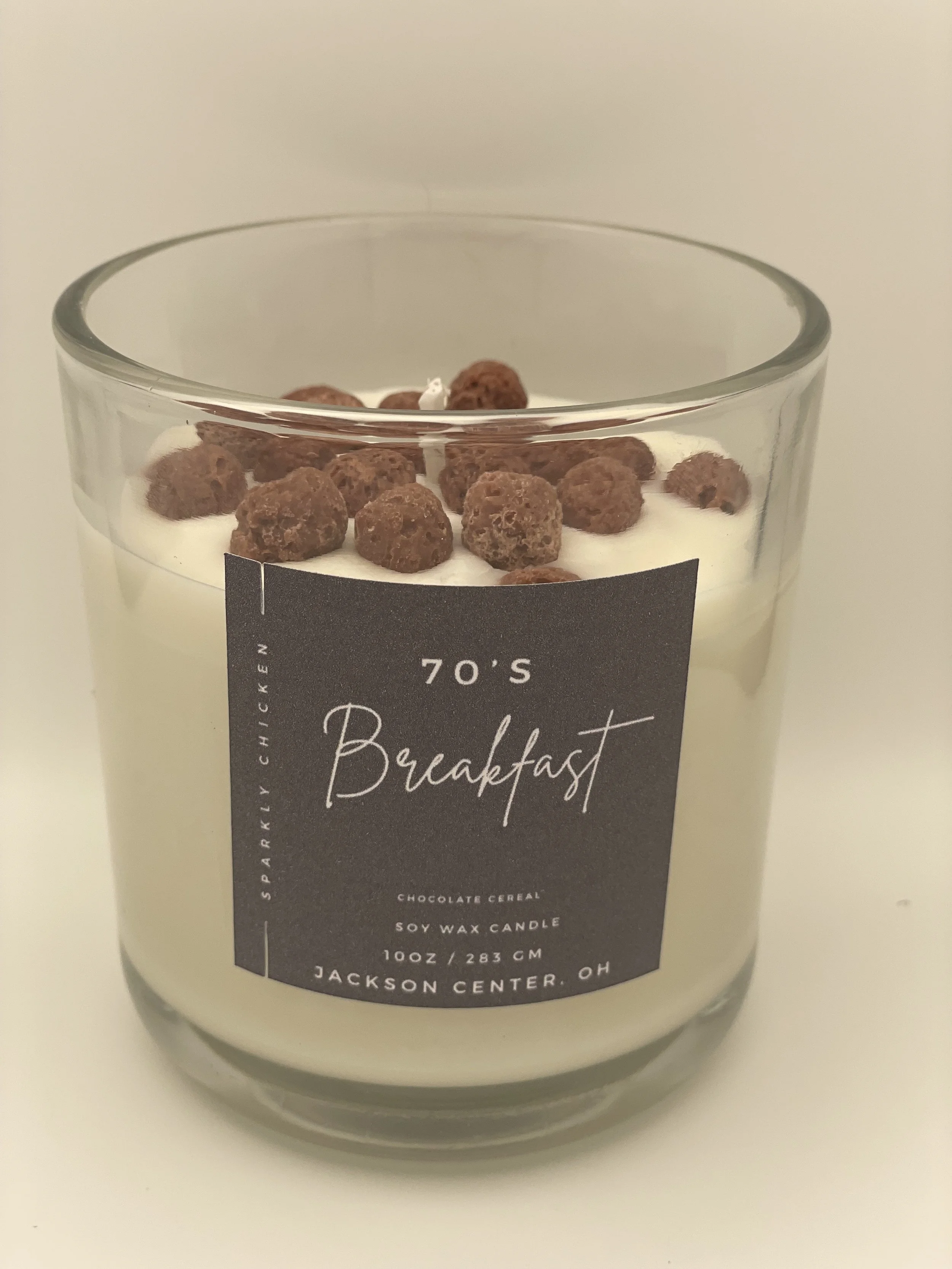 A candle in a glass holder labeled '70's Breakfast,' with chocolate cereal and soy wax.