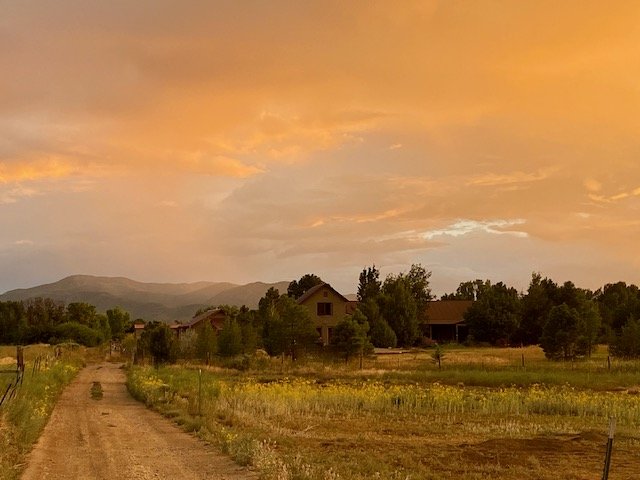 Rural dirt road leading to houses surrounded by fields and trees, with mountains in the background and a vibrant orange sunset sky overhead.