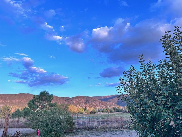 Blue sky with clouds over mountains and trees in a rural landscape.