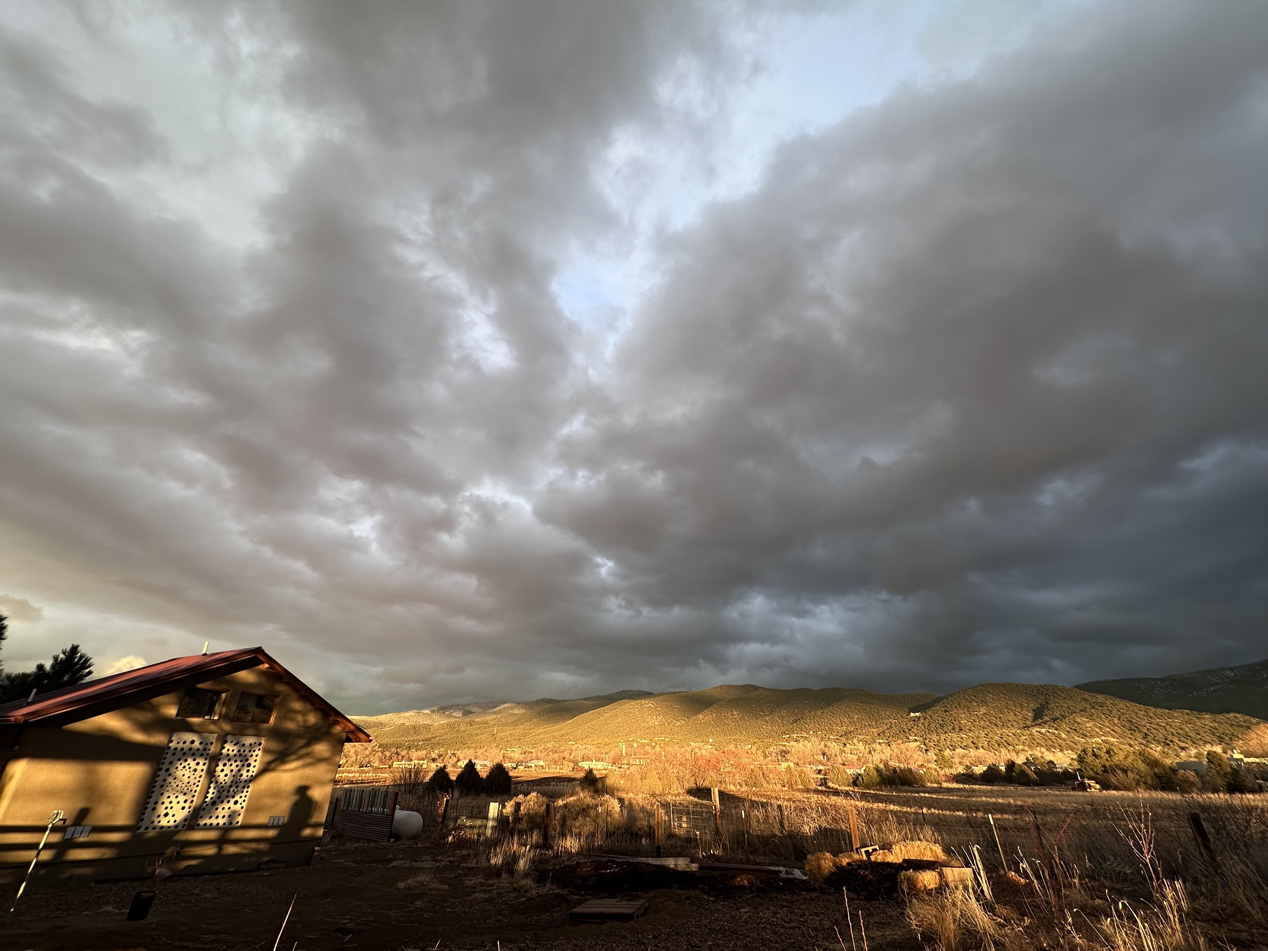 Landscape with dark cloudy sky, a small building to the left, sunlit hills in the background, and open fields with scattered bushes and fences.