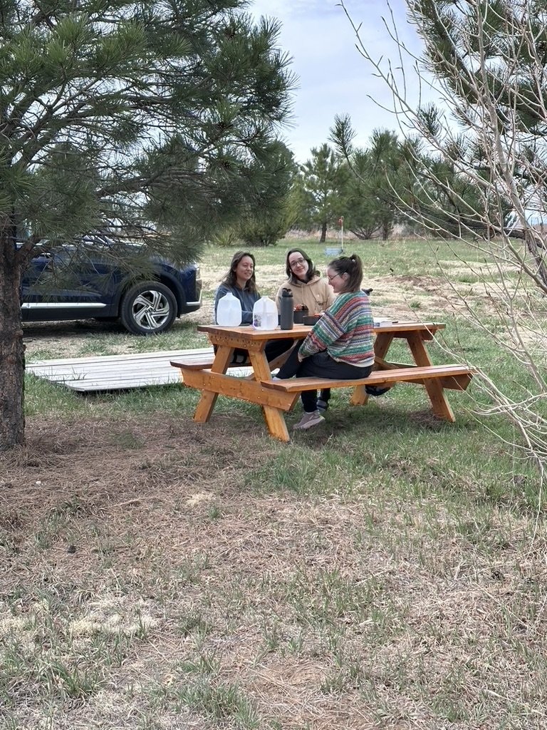 Three women sitting at a picnic table outdoors with water bottles and trees in the background.
