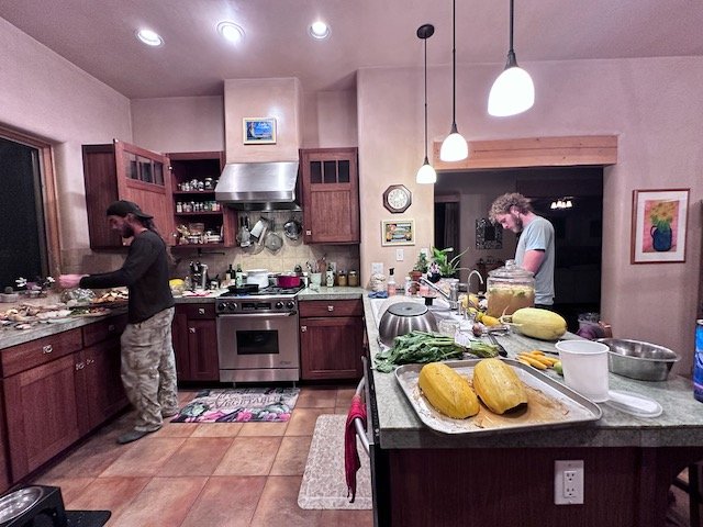 A modern kitchen with two people preparing food. The kitchen has wooden cabinets, a stainless steel stove, and a variety of kitchenware. Vegetables and kitchen tools are spread on the countertops, and pendant lights hang from the ceiling.