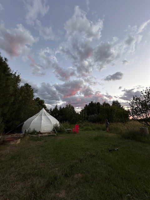 Outdoor scene at Harmony Farm, Taos New Mexico with a glamp tent on a grassy field, surrounded by trees, with a red chair nearby and a person standing in the distance under a cloudy sky at dusk.
