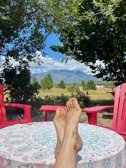Feet resting on a table with a pink floral tablecloth, surrounded by pink chairs, with a scenic view of trees, Pueblo mountain, Taos, and a blue sky in the background.