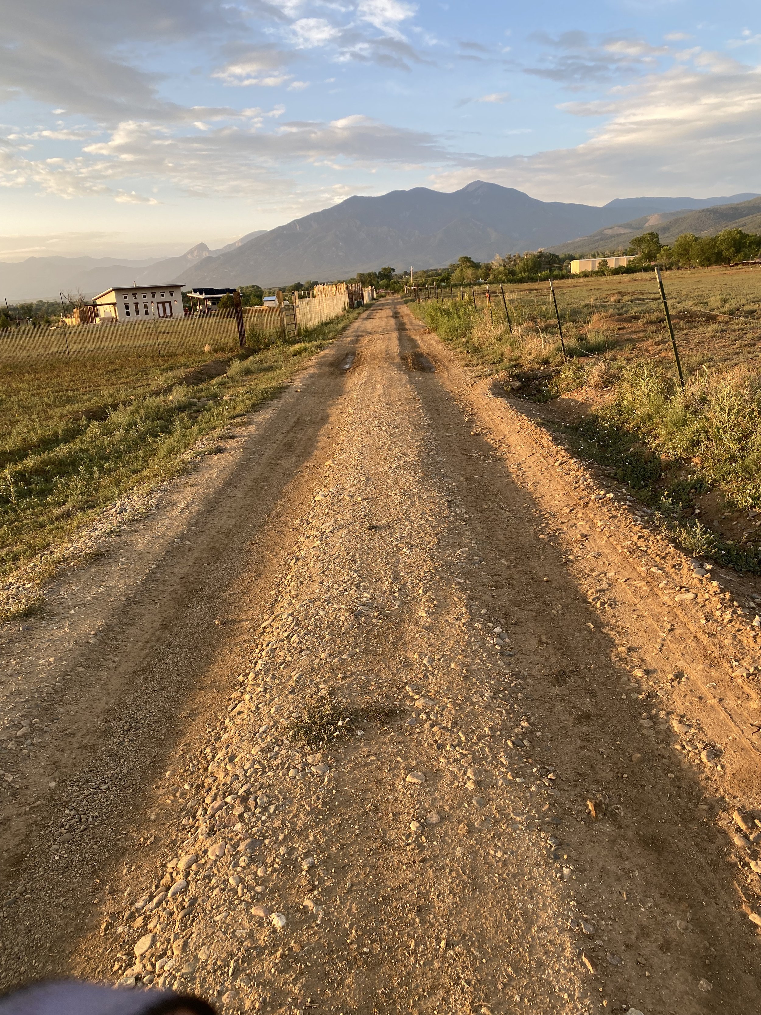 Dirt road leading towards mountains, flanked by fields and fences, with buildings in the distance during sunset.