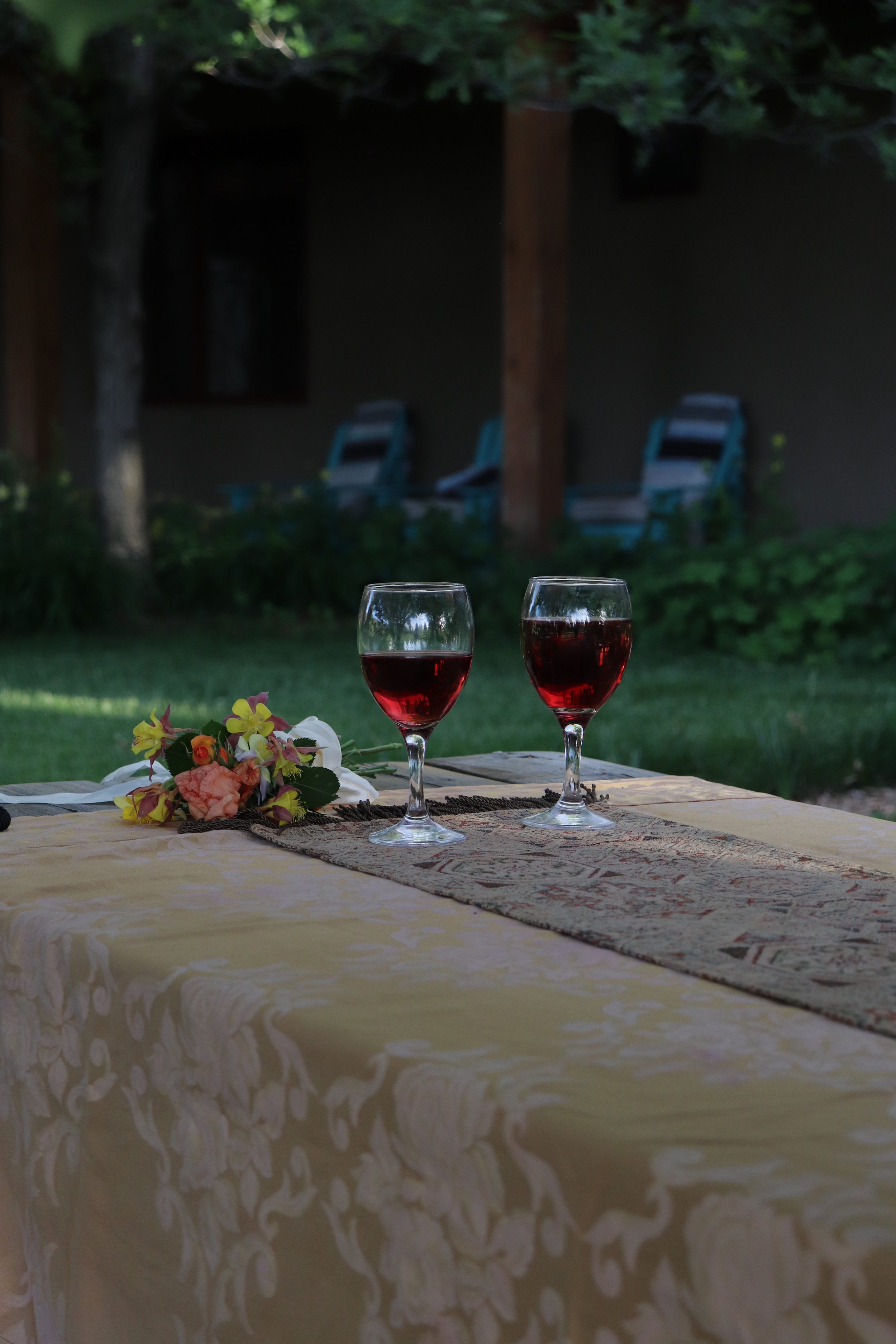 Two glasses of red wine on a wedding picnic table with a floral tablecloth and a bouquet of flowers, outdoor setting.