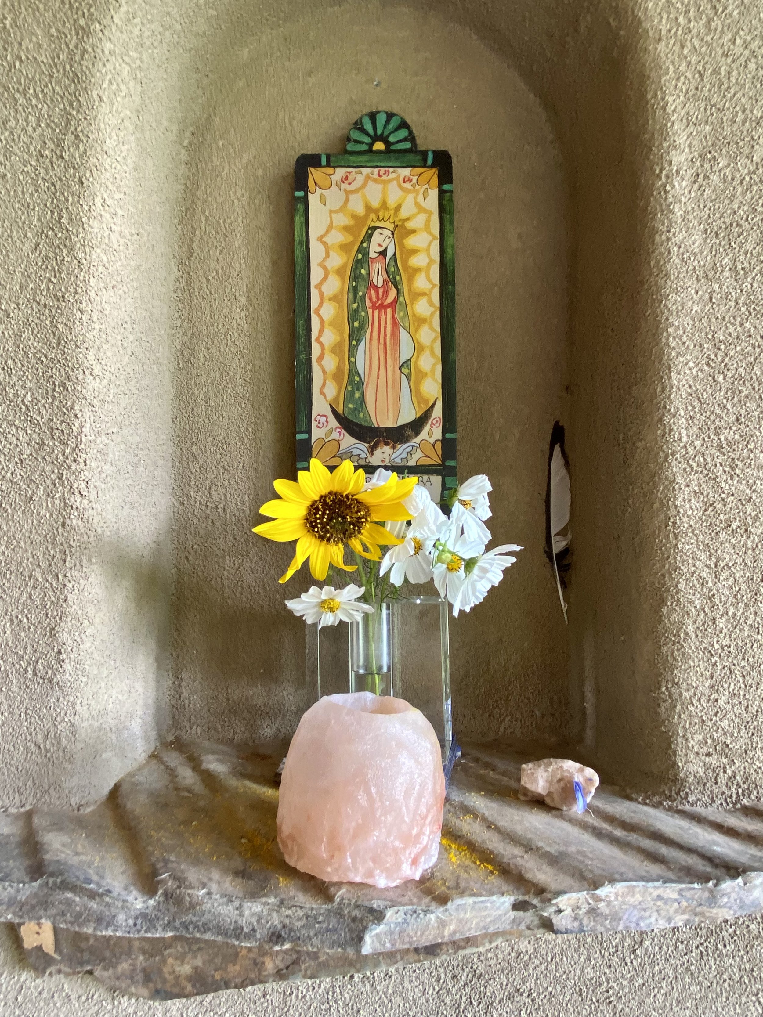 A decorative alcove with a Patron saint, Guadalupe , a sunflower and white flowers in a glass vase, a pink Himalayan salt candle holder, and a small crystal on a stone shelf.