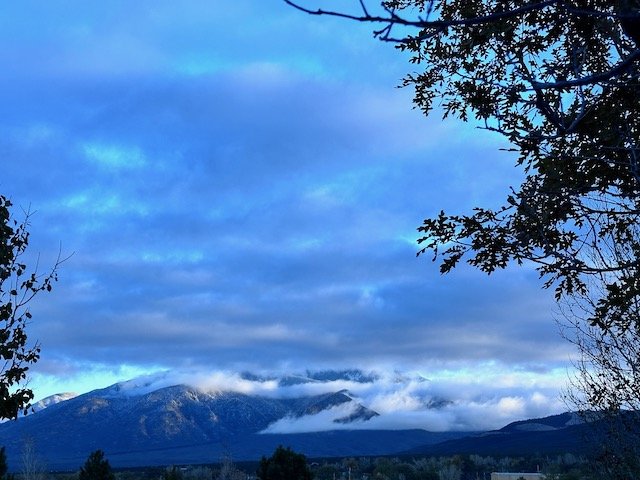 Mountains covered in clouds under a blue sky with tree branches framing the scene.